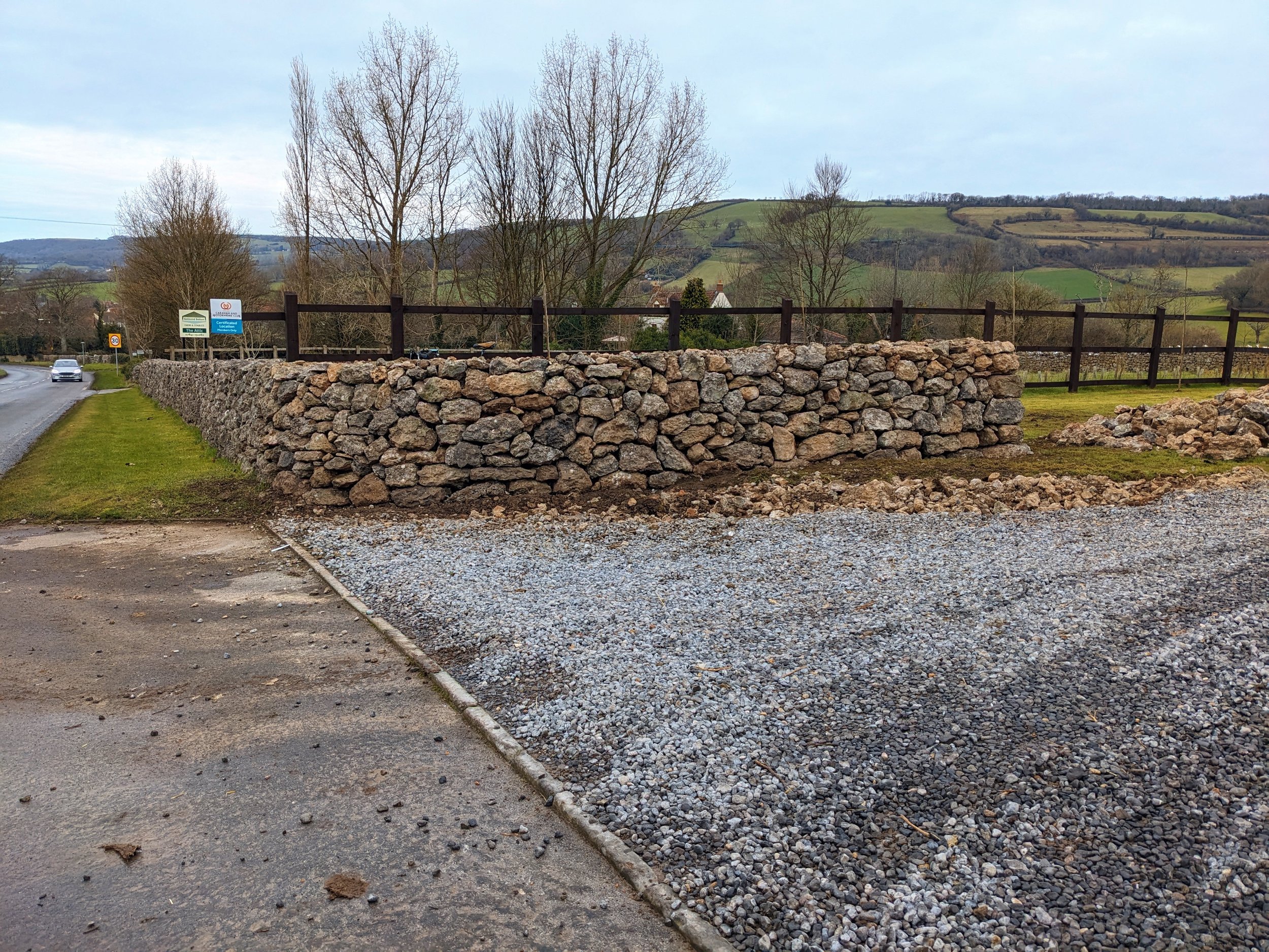 A dry stone wall entrance in Easton, Somerset with a wooden fence on top, beside a road with a car, in a rural landscape with trees and green hills in the background.