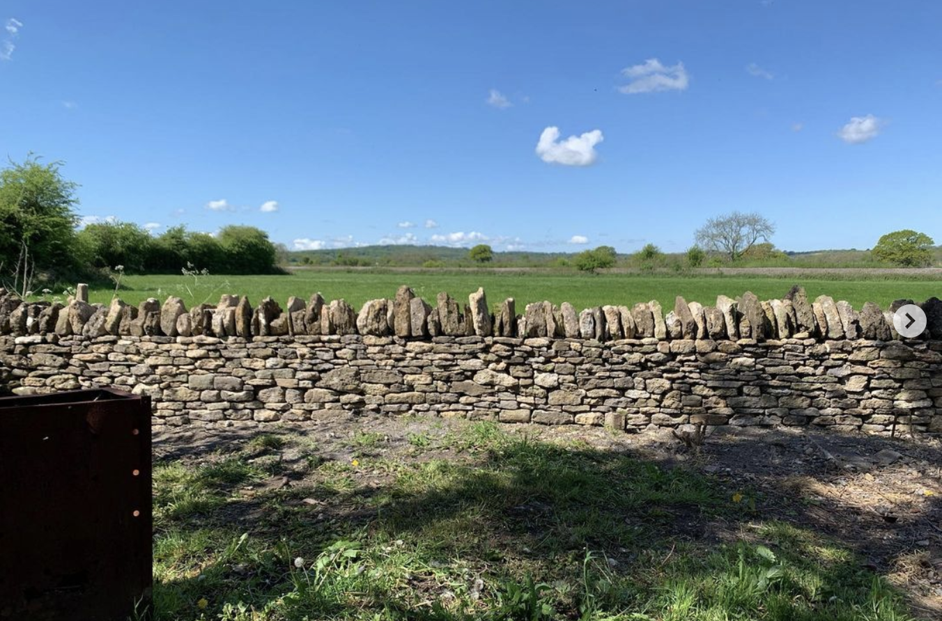 A dry stone wall in Frome set in a rural landscape under a blue sky with scattered clouds, green fields, and distant trees.