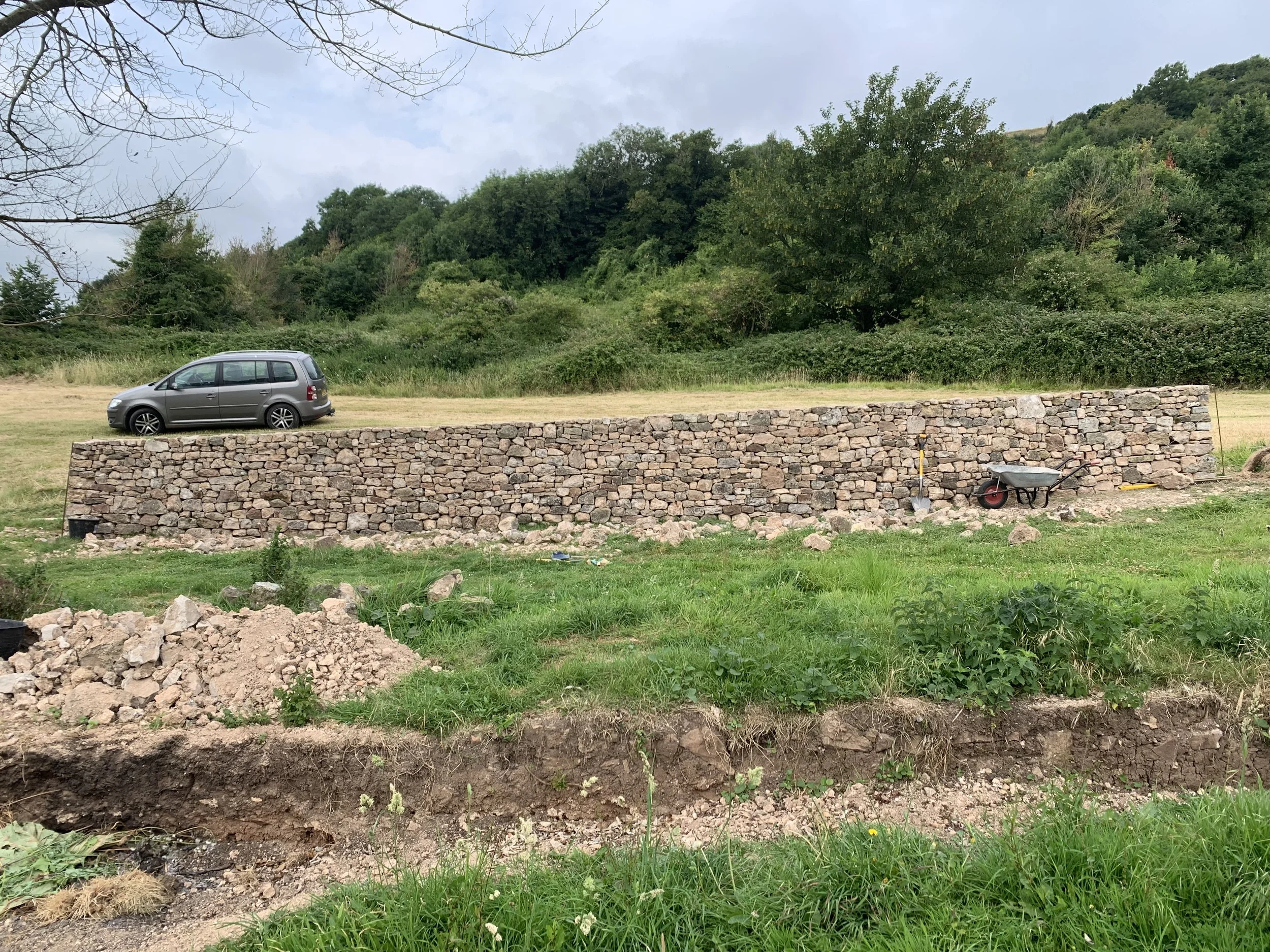 A dry stone wall in Compton Bishop, Somerset with a wheelbarrow and a shovel resting against it. Behind the wall, a gray minivan is parked on a grassy area with trees and a hillside in the background.