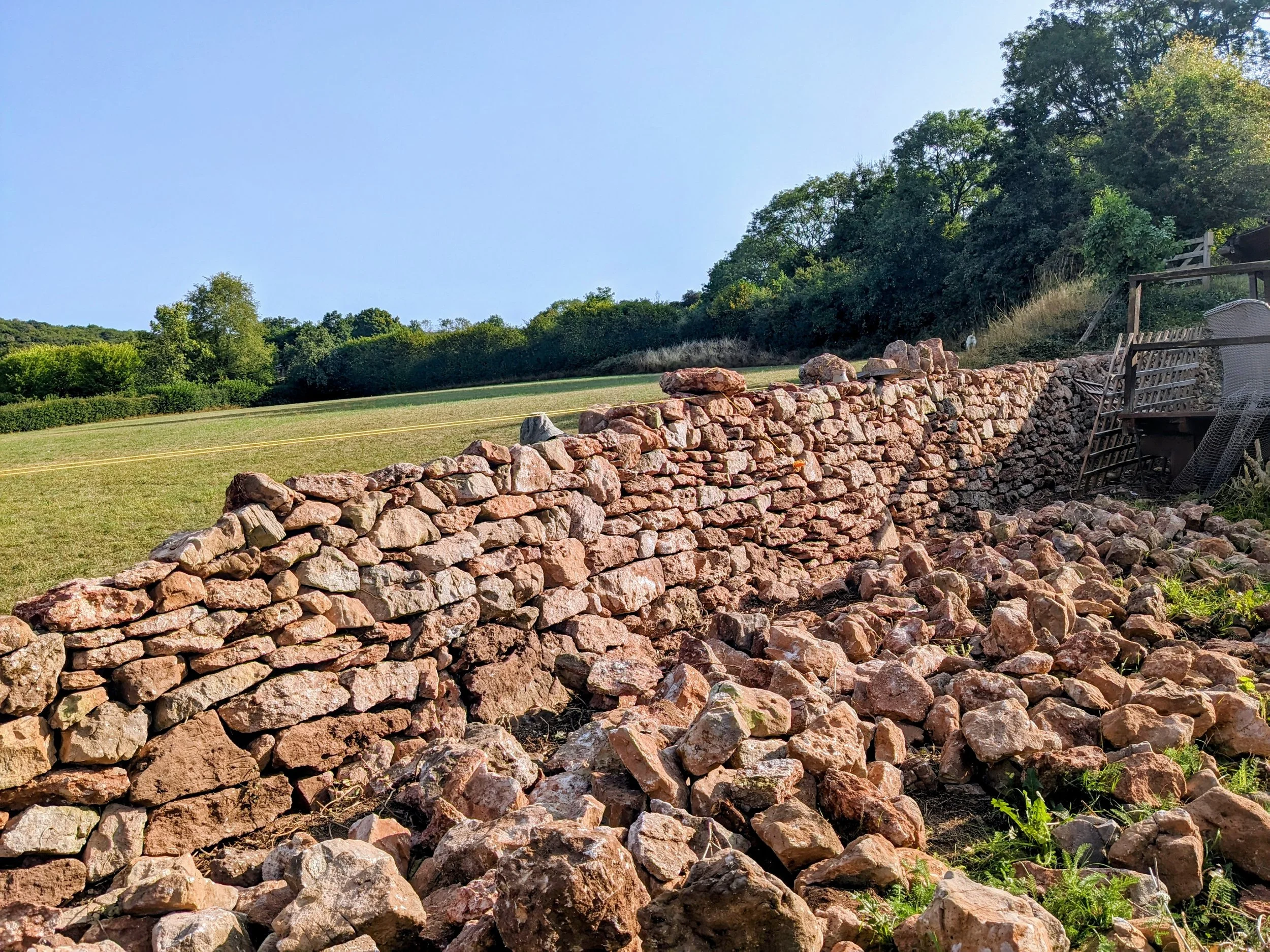 A dry stone wall in Cheddar, Somerset build in a random rubble style, set against a backdrop of green trees and a clear blue sky, with outdoor furniture and a lattice on the right side.
