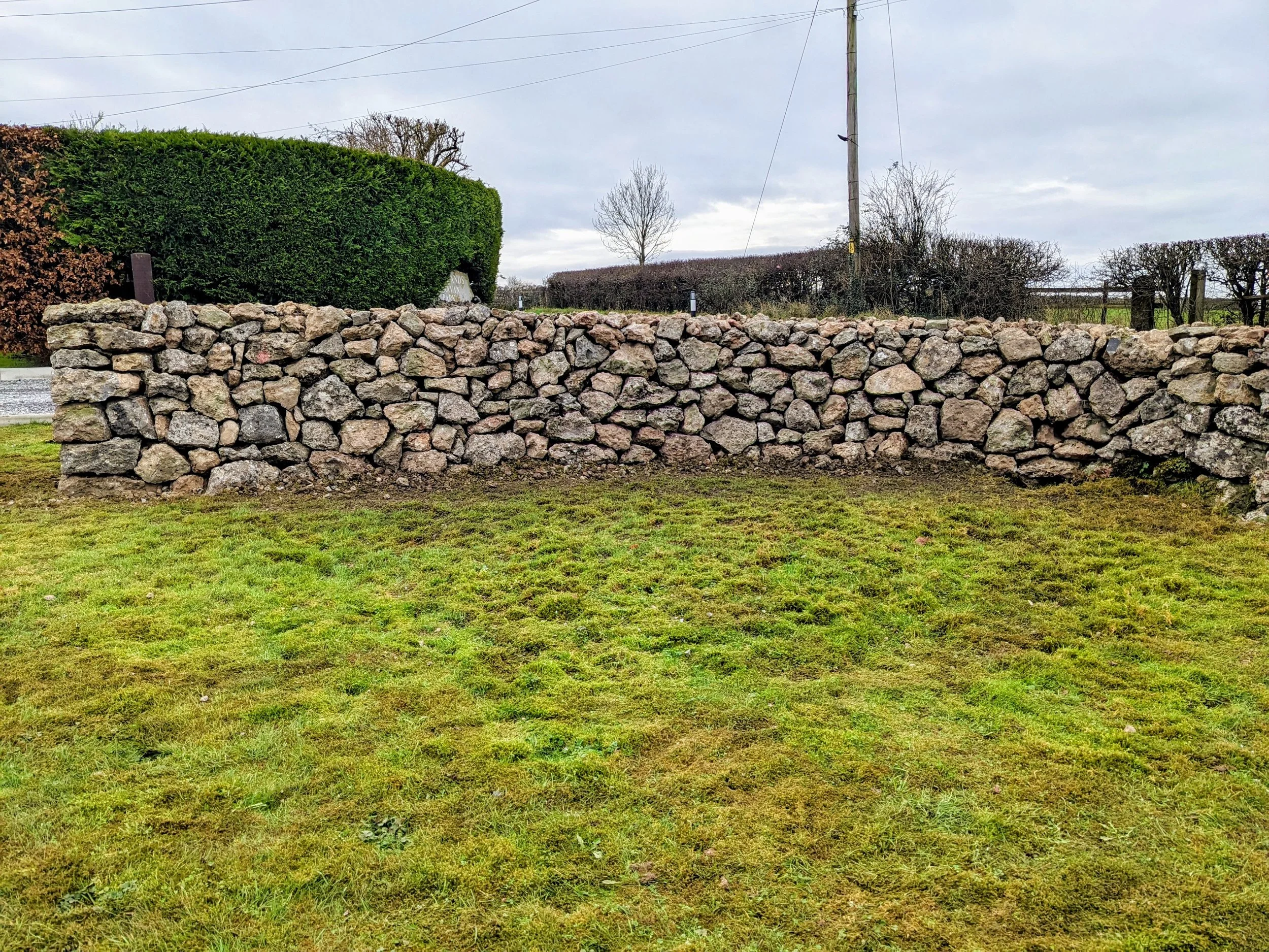 A dry stone wall in Easton, Somerset running horizontally across a grassy yard, with bushes and trees in the background under a cloudy sky.