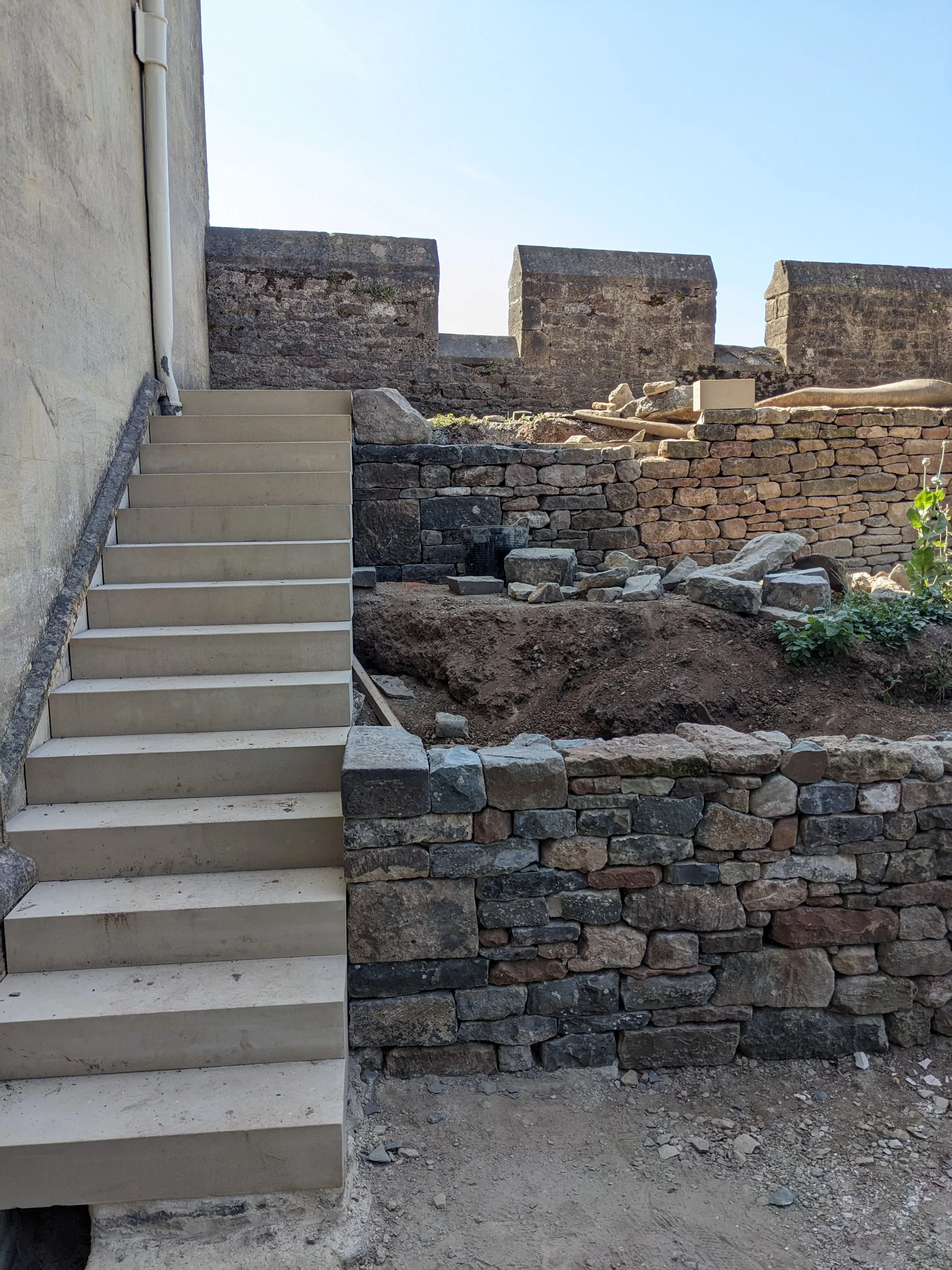 Stone stairs next to a wall under construction, with a layered stone retaining wall in the background, and some dirt and rocks.