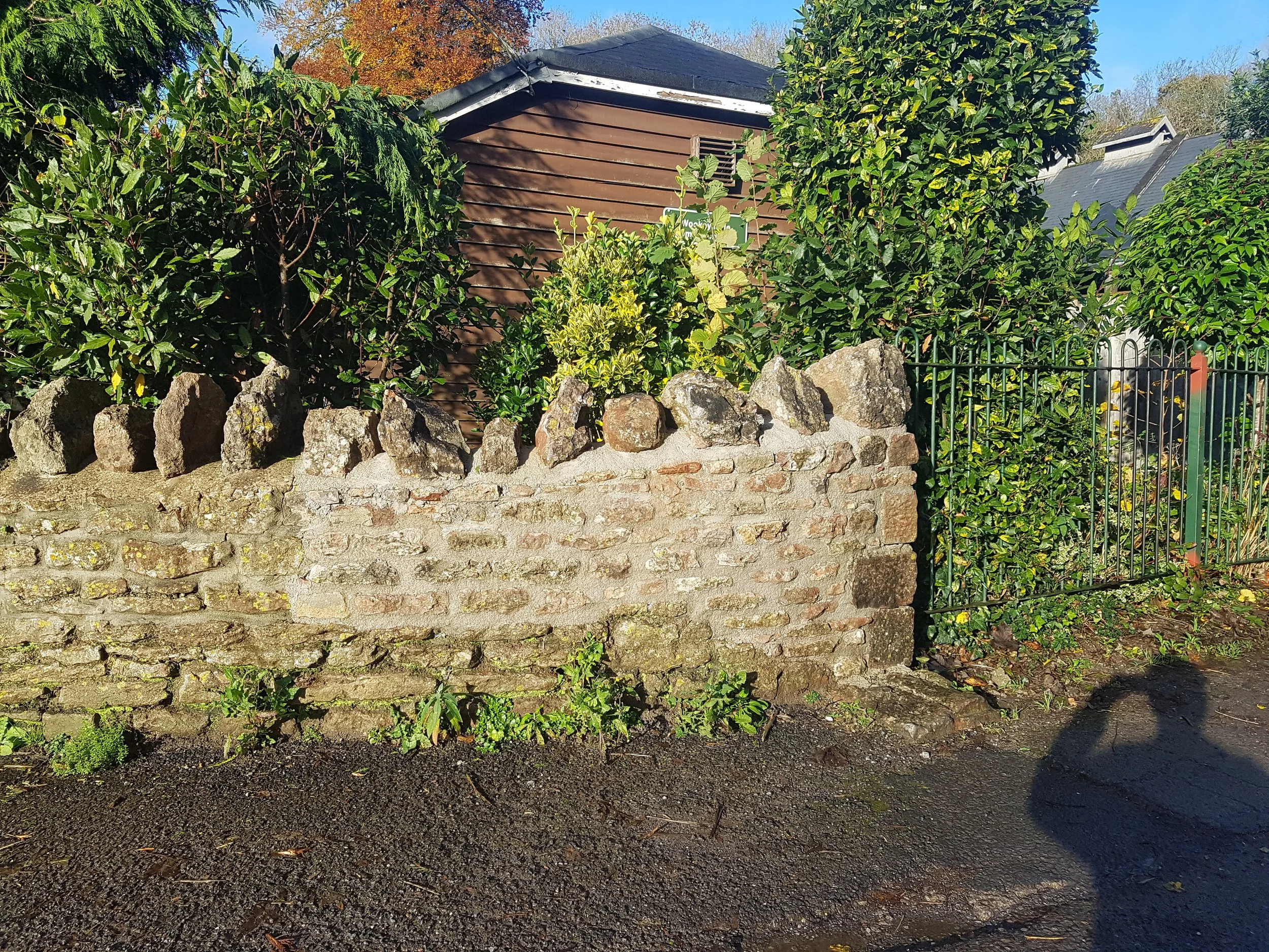 A lime mortared stone wall in Wookey Hole, Somerset with greenery and a brown wooden building in the background, a black metal fence on the right, and a person's shadow on the ground in the foreground.