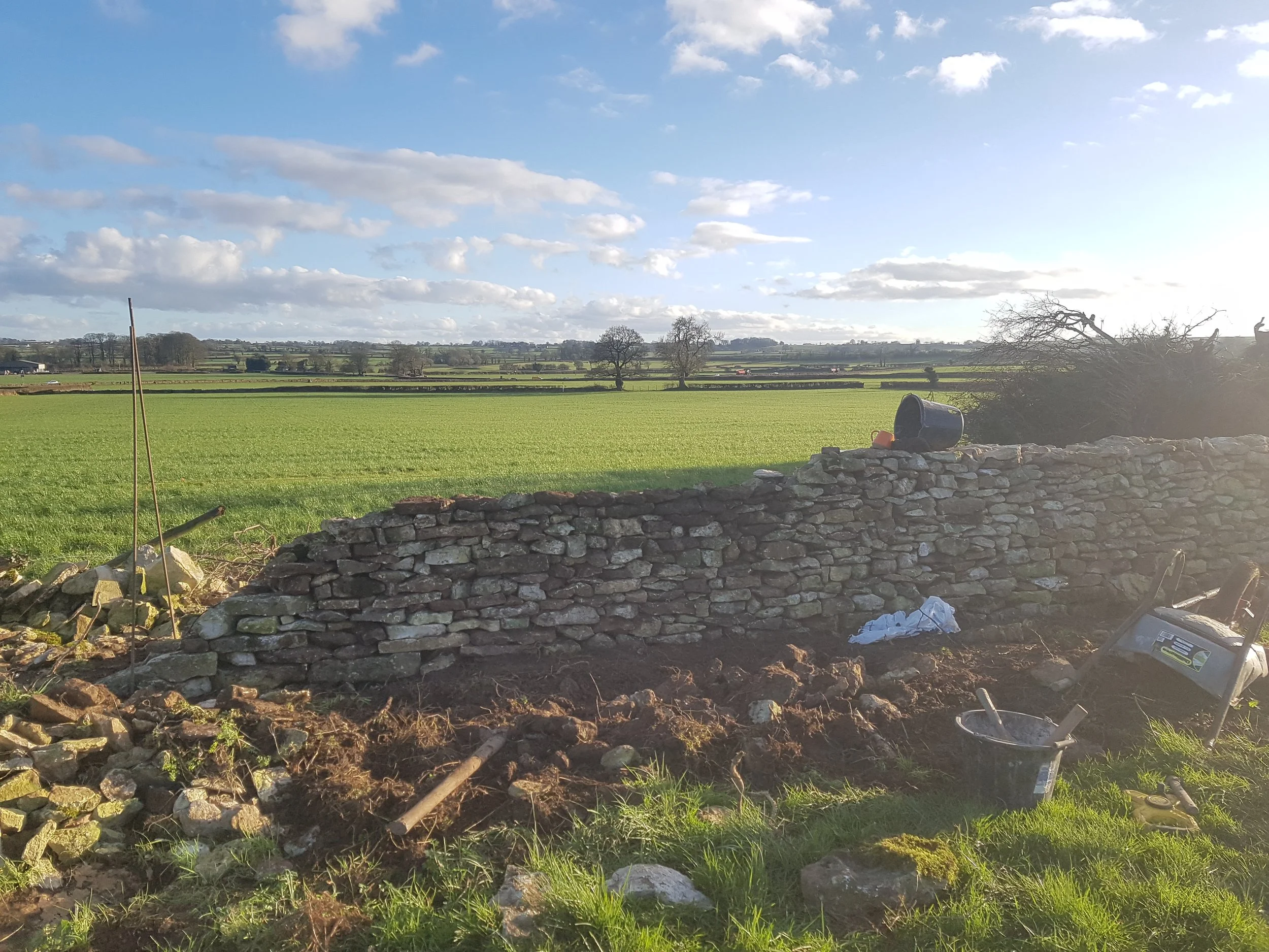 A dry stone wall in Chewton Mendip, Somerset under construction in a rural landscape with green fields, trees, and a partly cloudy sky in the background. Construction tools and materials are visible nearby.