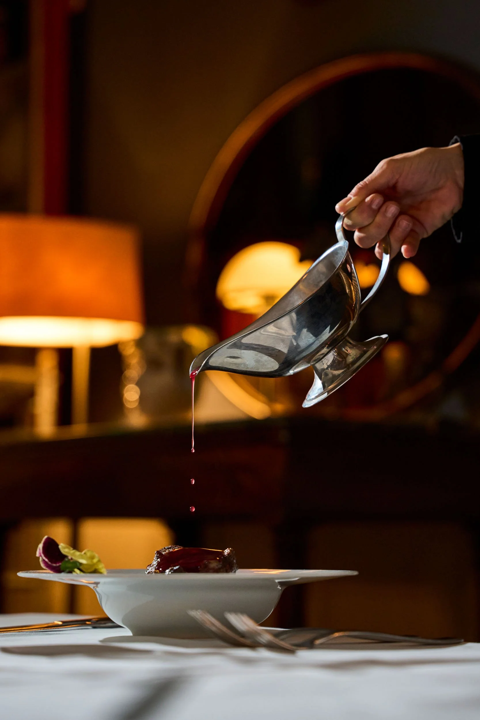 Un uomo versa una salsa da un cucchiaio su un piatto bianco con un pezzo di carne e decorazioni da insalata, in un ambiente elegante con luci calde.