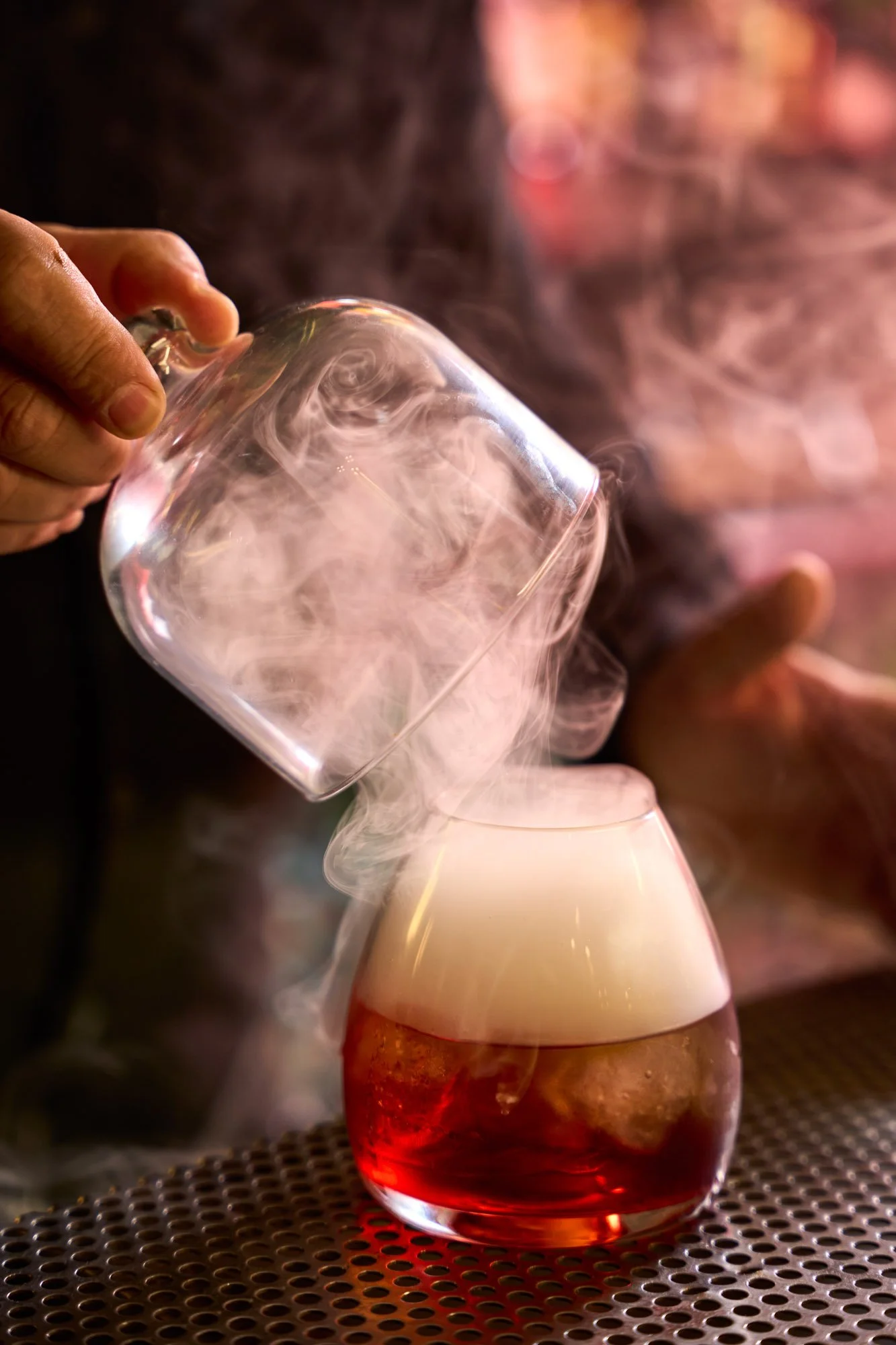 Una mano versa un liquido colorato in un bicchiere di vetro con vapore o fumo che sale, creando un effetto visivo di fumo o nebbia sopra il bicchiere.