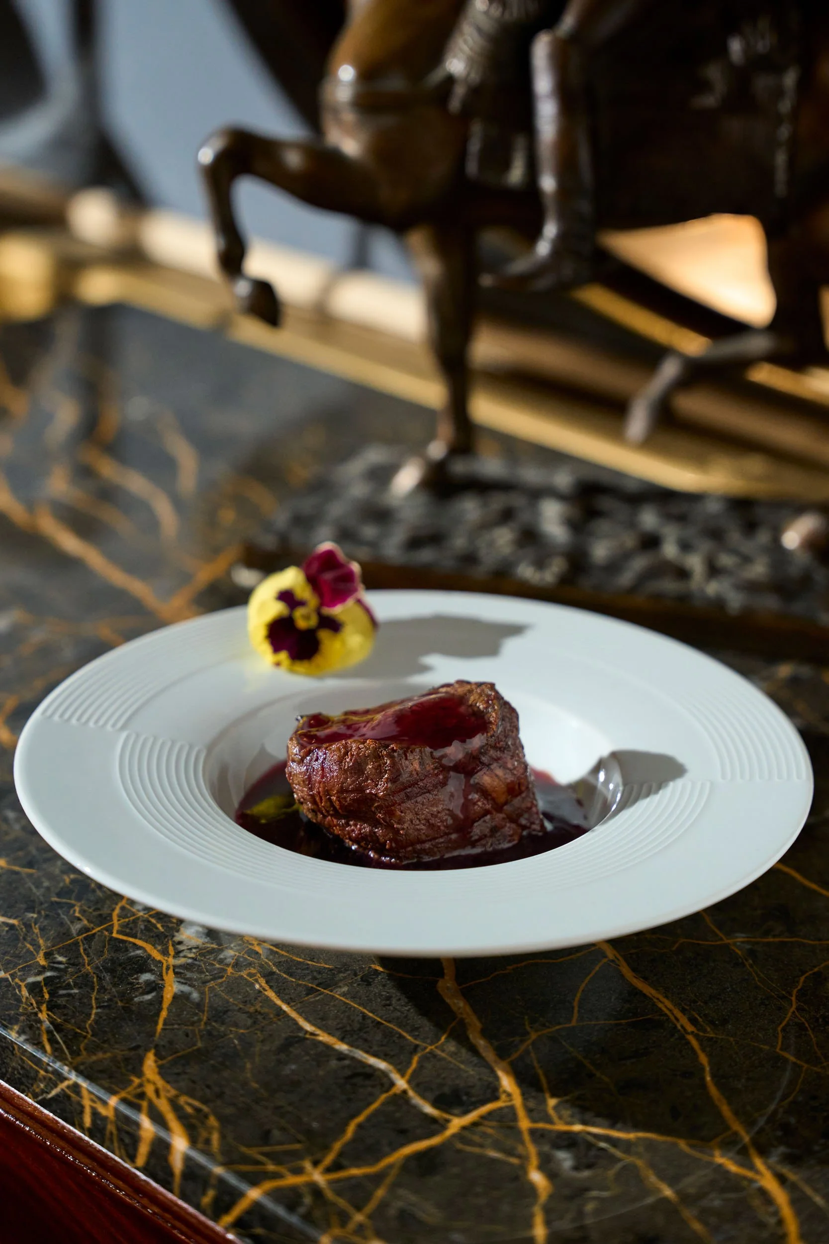Una fetta di carne in umido con salsa, accompagnata da una decorazione floreale viola e gialla su un piatto bianco, posato su un tavolo in marmo scuro con venature dorate.