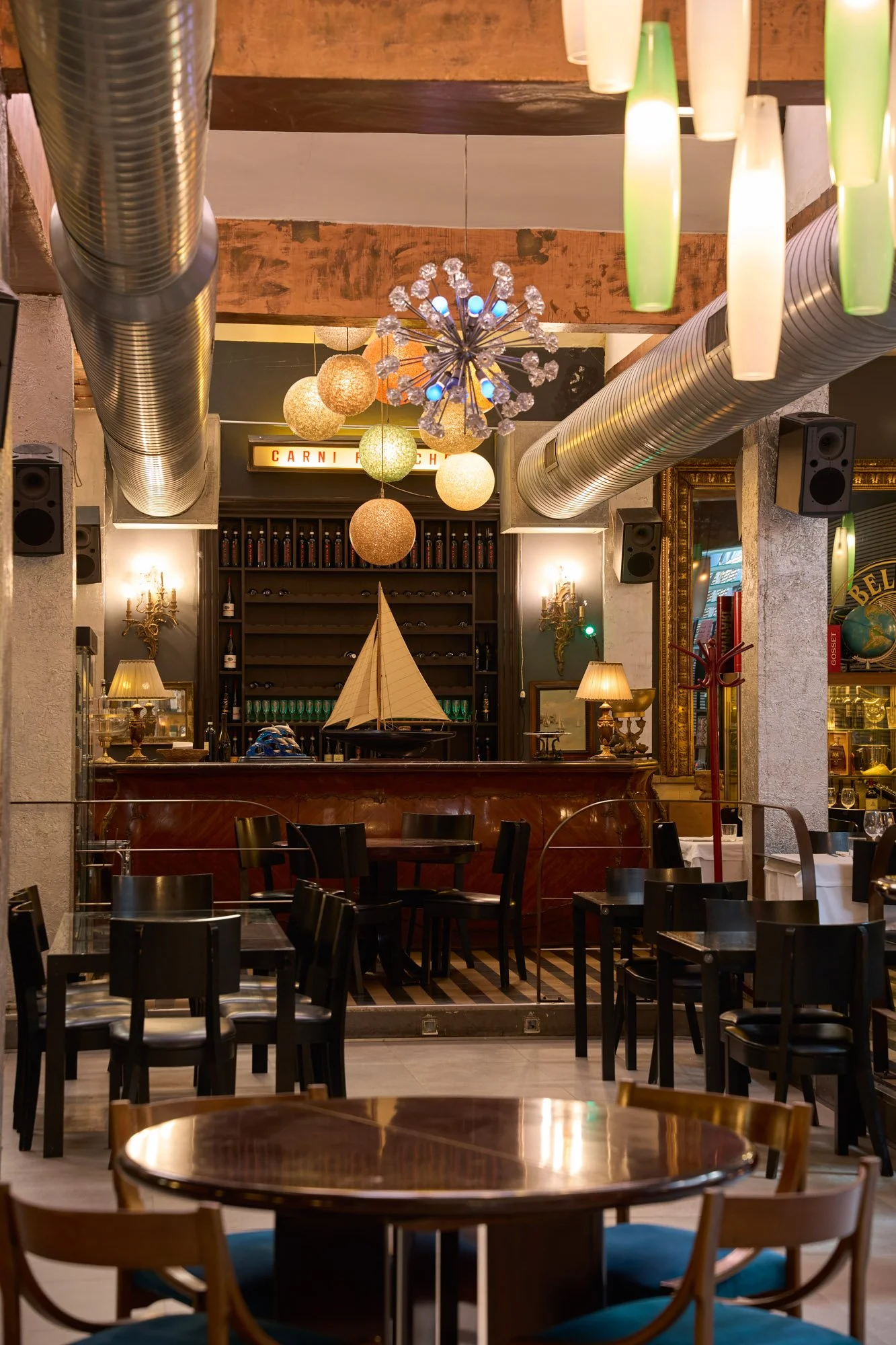 Interno di un ristorante con tavoli e sedie in primo piano, decorazioni moderne e vintage, illuminazione calda, lampade di diverse forme e colori, ripiano con bottiglie e decorazioni, bar con modello di nave in miniatura, specchio e quadri alle pareti.