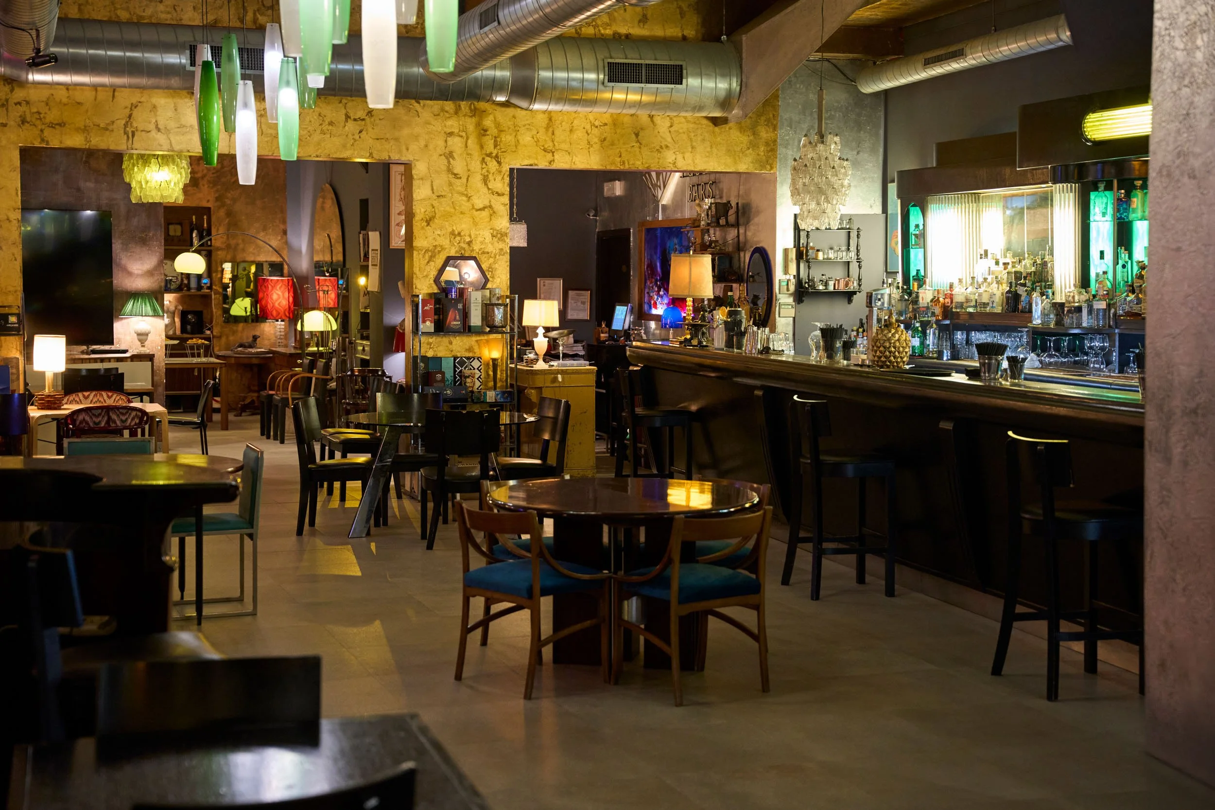 Interno di un bar con tavoli, sedie e una zona bar con bottiglie e illuminazione colorata.