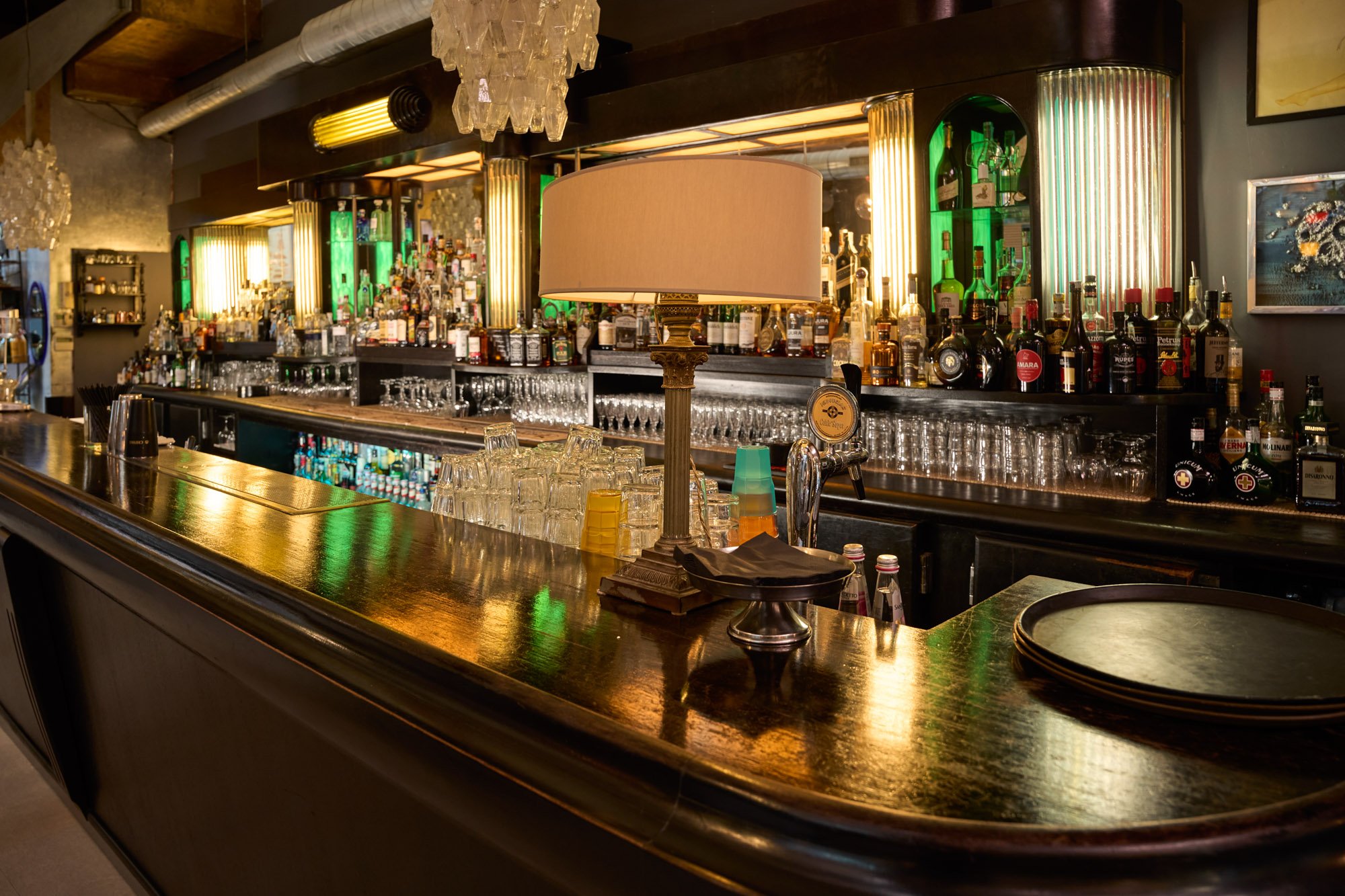 Bancone di un bar elegante con bicchieri, bottiglie di alcolici sullo sfondo, una lampada decorativa e stoviglie nere.