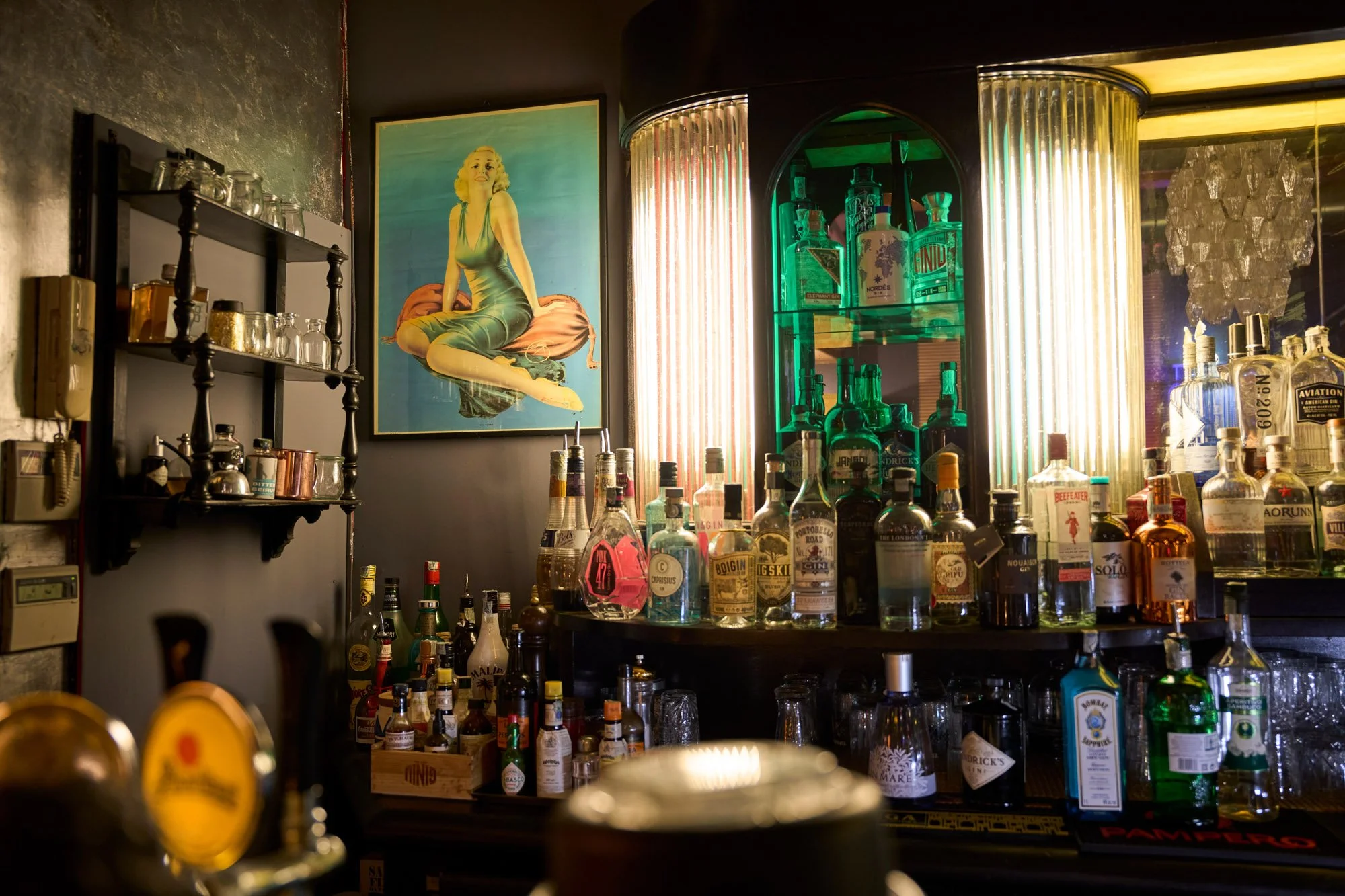 Interno di un bar con bottiglie di alcolici su uno scaffale e al centro un bancone con bottiglie, in basso un tappetino con la scritta 'DAMPFER'. Sul muro una stampa di una donna sorridente in posa seduta, e alle spalle un'illuminazione con luci colorate.