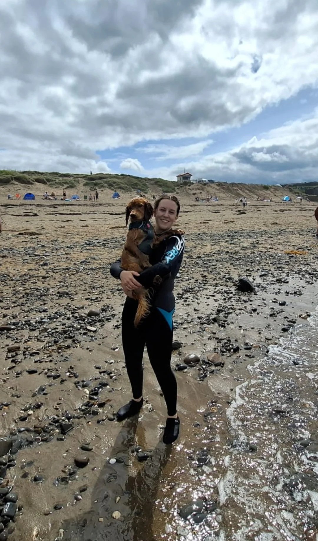 A woman in a black wetsuit standing on a rocky beach, holding a dog in her arms. The sky is cloudy, and there are people and tents in the background.