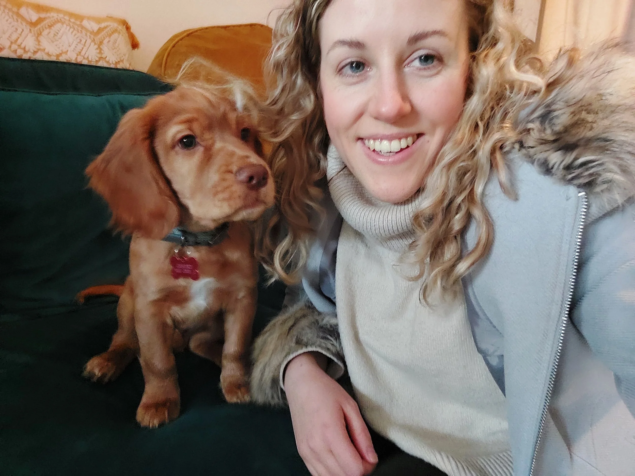 A smiling woman with curly blonde hair taking a selfie with a brown puppy sitting next to her on a dark green sofa.