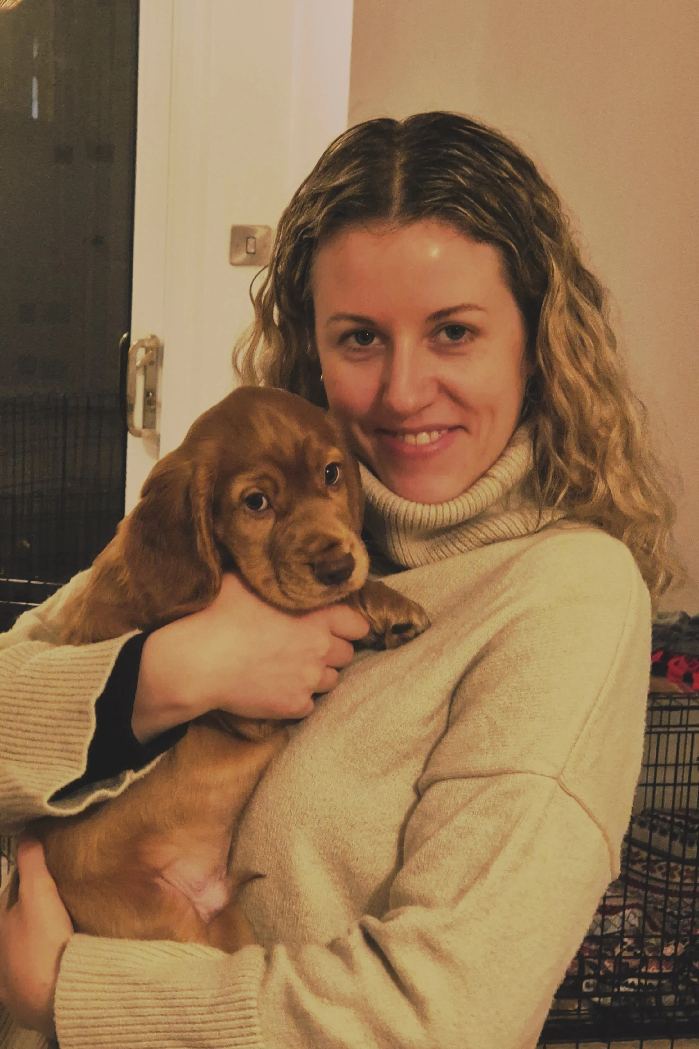 A woman with curly blonde hair holding a brown puppy inside a home.