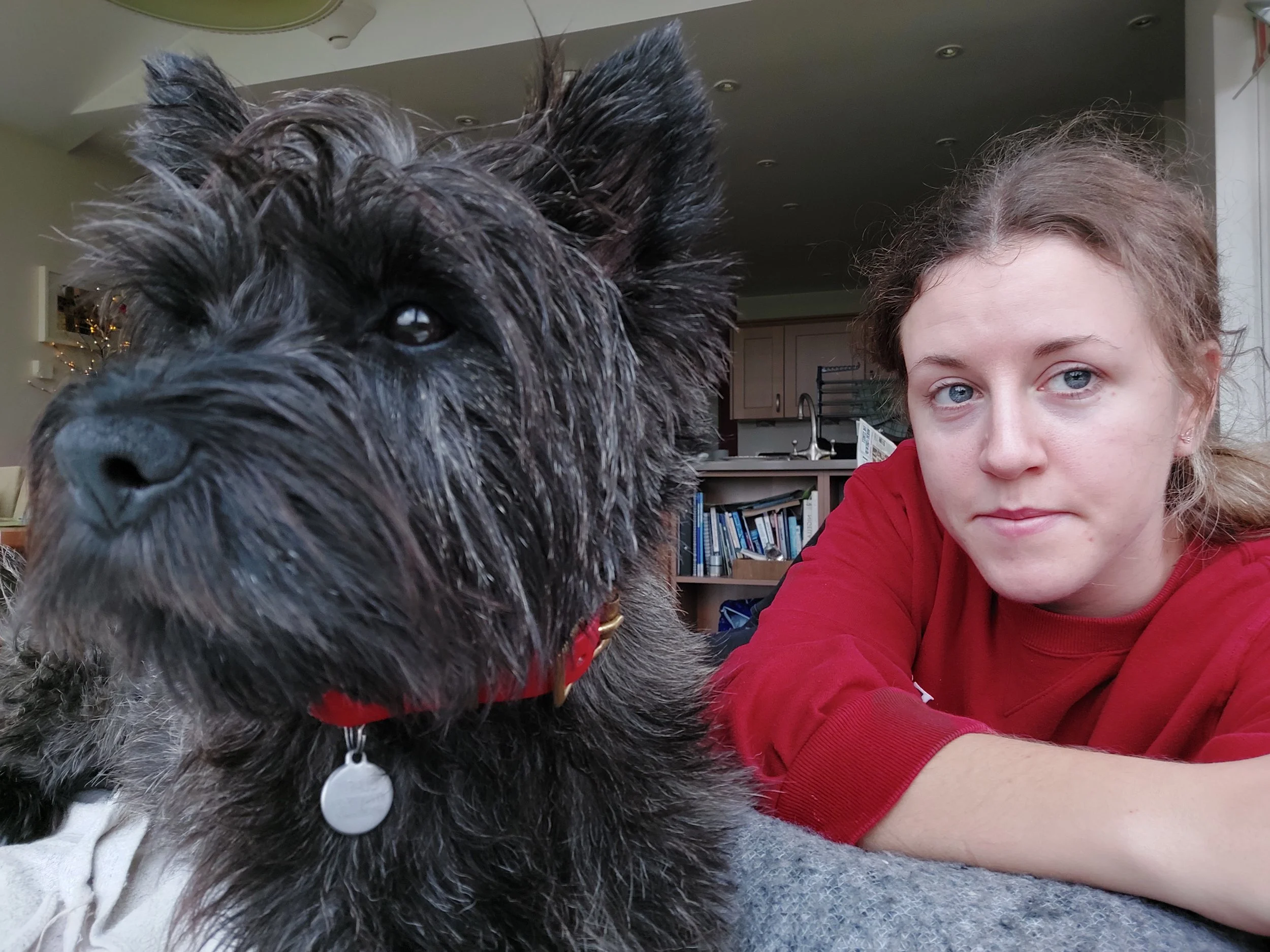 A woman and a large black dog with gray fur on its face, lying on a gray surface indoors. The woman has light skin, curly brown hair, and blue eyes, wearing a red long-sleeve shirt. The background shows a kitchen with beige cabinets, a sink, and book