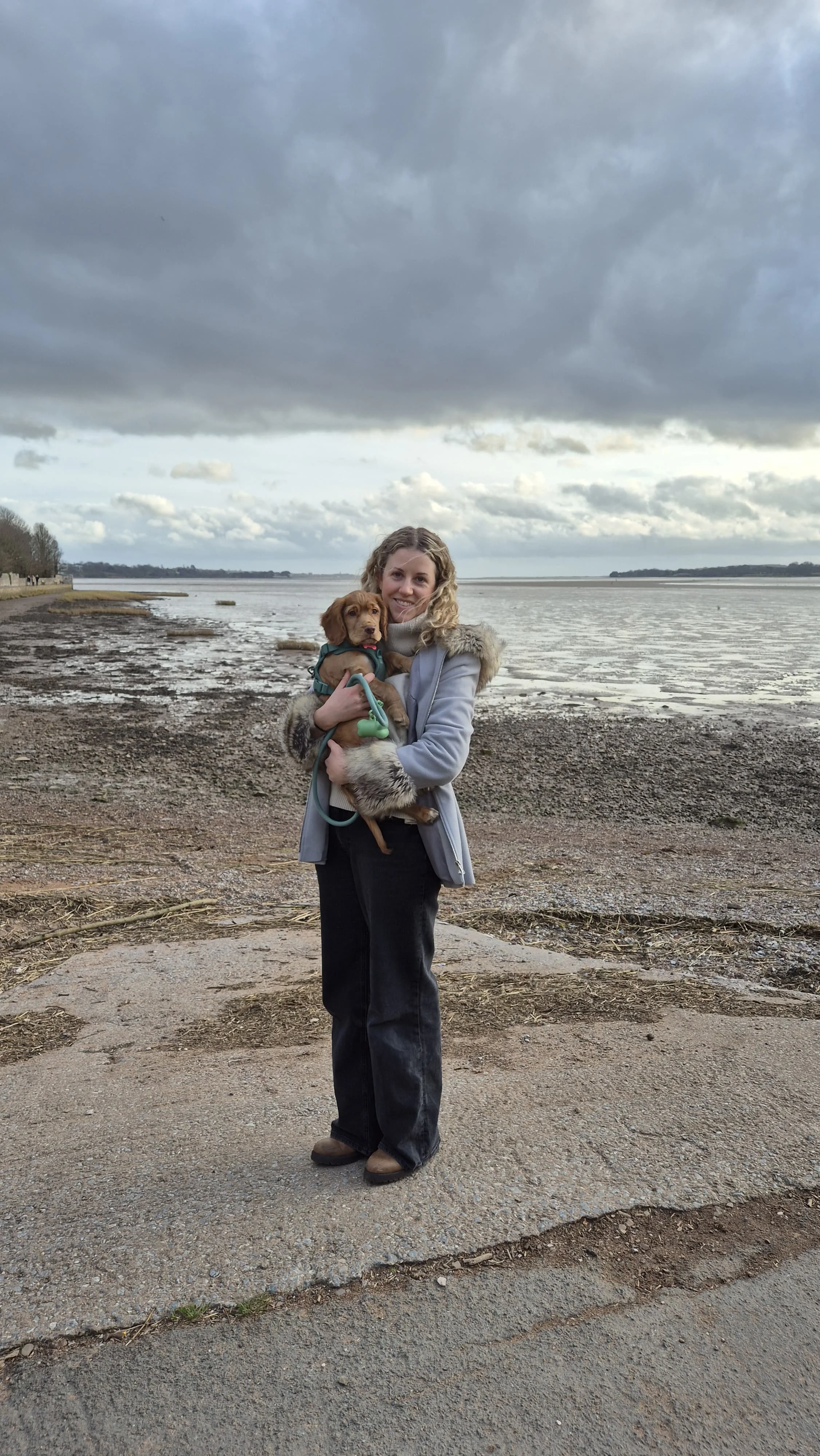 A woman holding a puppy by a body of water under a cloudy sky.