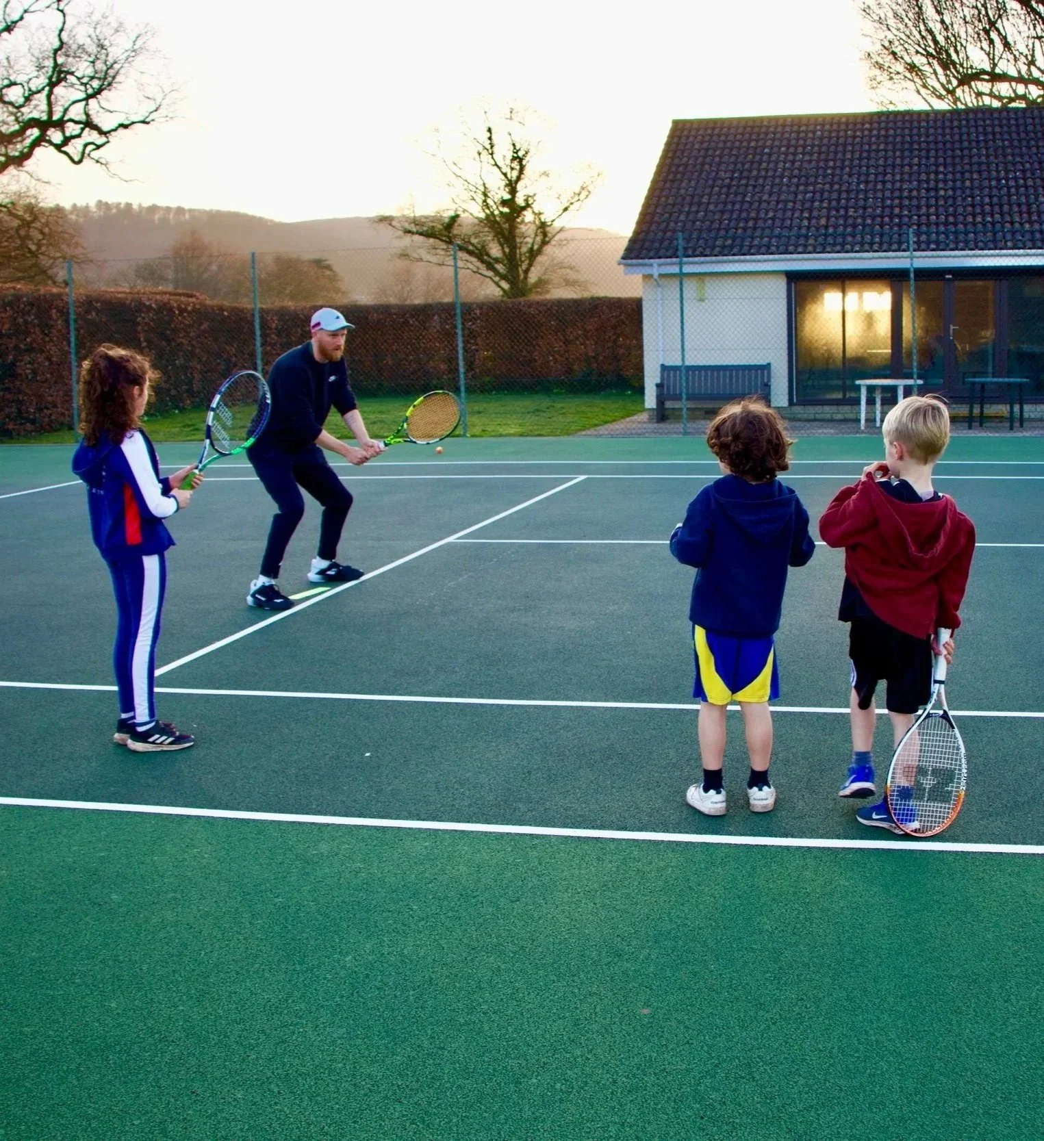 A man demonstrating tennis skills to three children on an outdoor tennis court during sunset.
