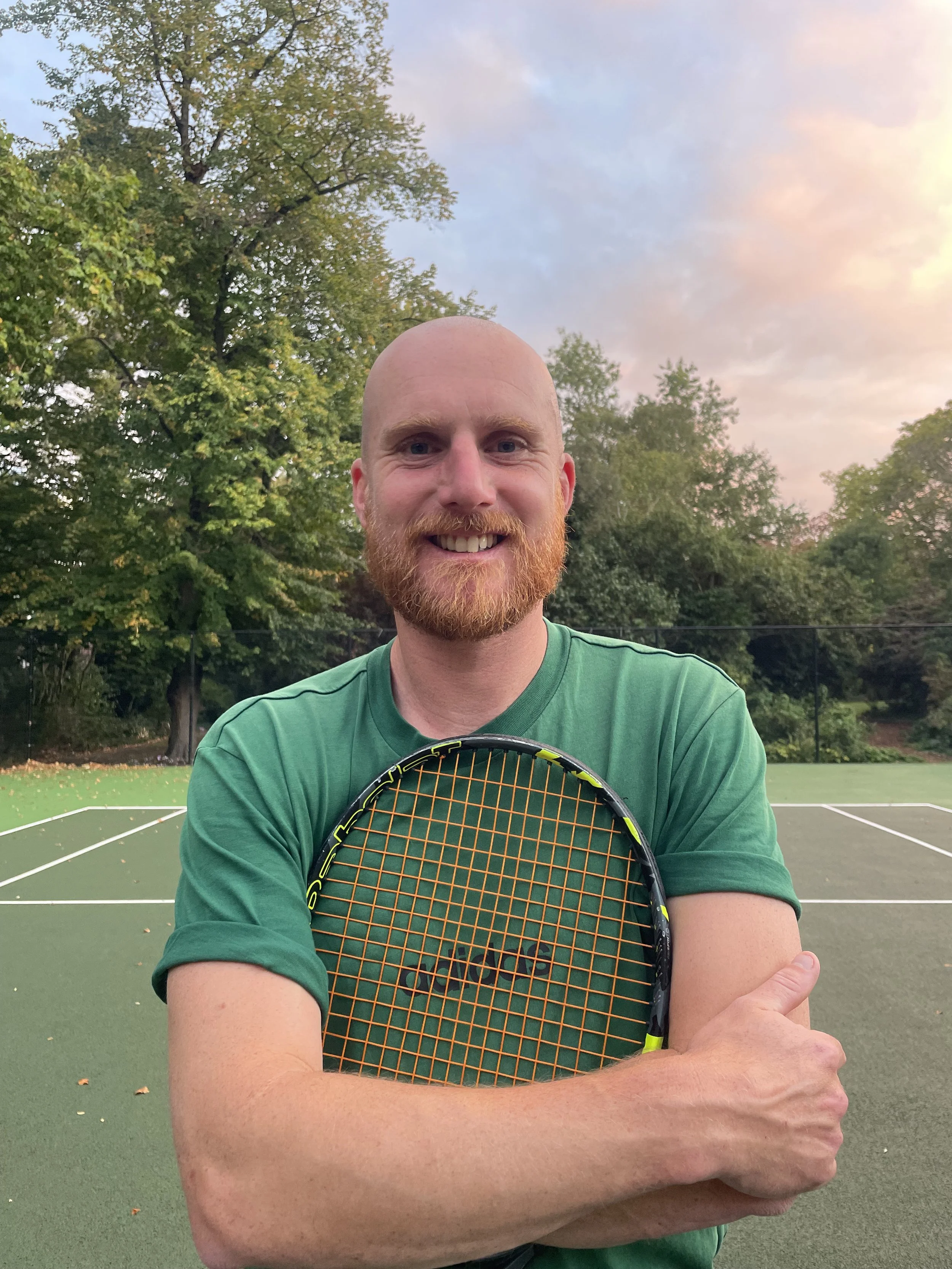 A man with a bald head and red beard holding a tennis racket on an outdoor tennis court during sunset with trees in the background.
