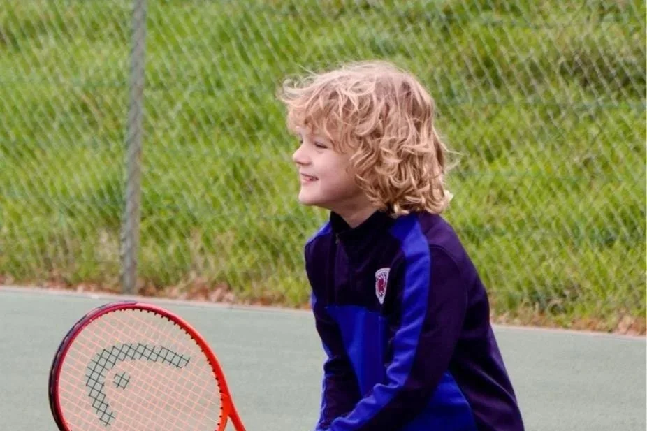 Child playing tennis on an outdoor court with a green grass backdrop and chain-link fence.