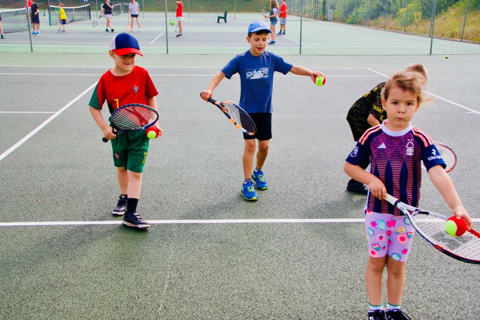 Four children standing on a tennis court holding tennis rackets and balls, with other kids playing in the background.