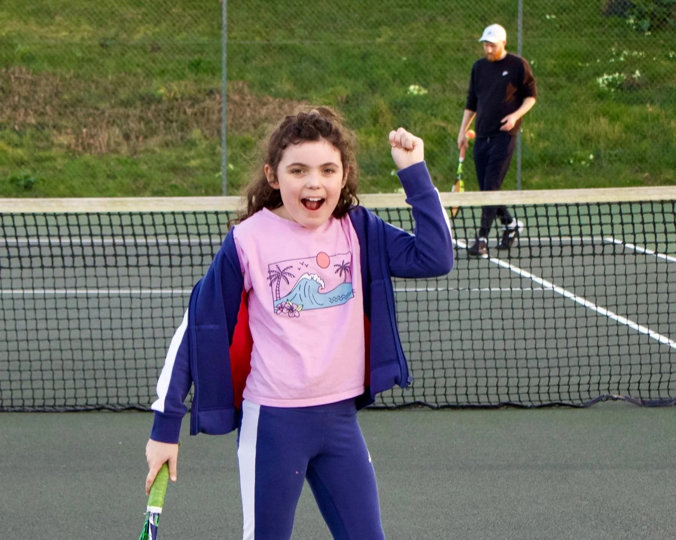 A young girl celebrating on a tennis court, holding a tennis racket, with a man in the background in a black and white outfit.