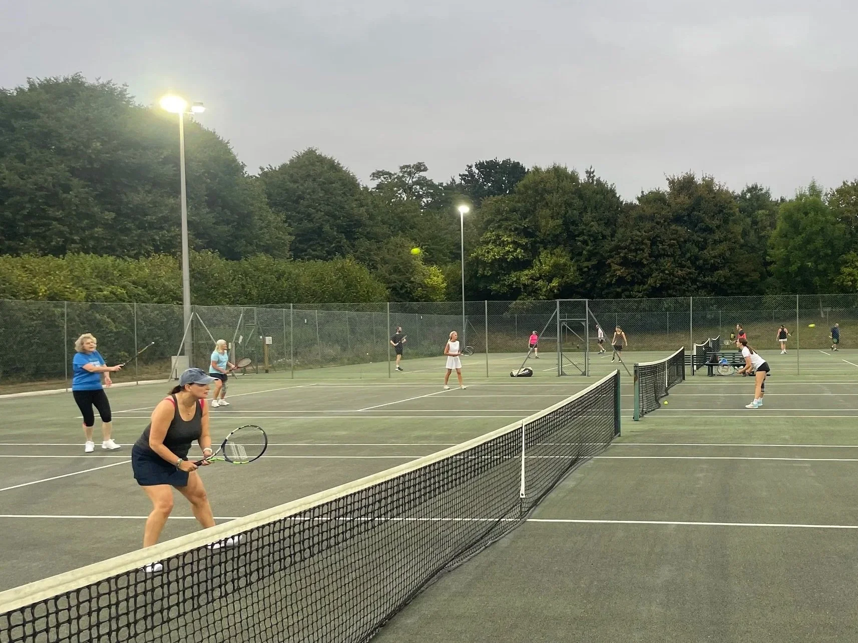 People playing tennis on multiple courts during overcast weather, with trees in the background and lit tennis courts.
