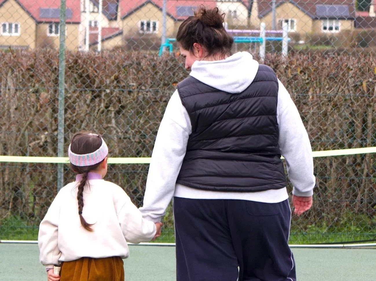 A woman and a young girl holding hands on a tennis court, with the woman looking down at the girl, and a fence with bushes and houses in the background.