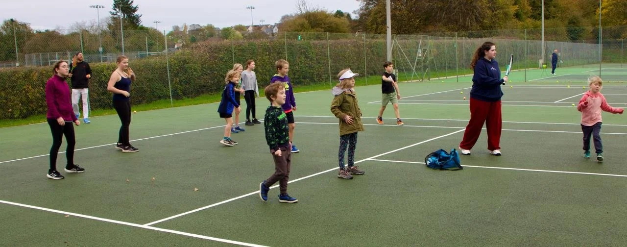 Children and a coach practicing tennis on an outdoor court during daytime.