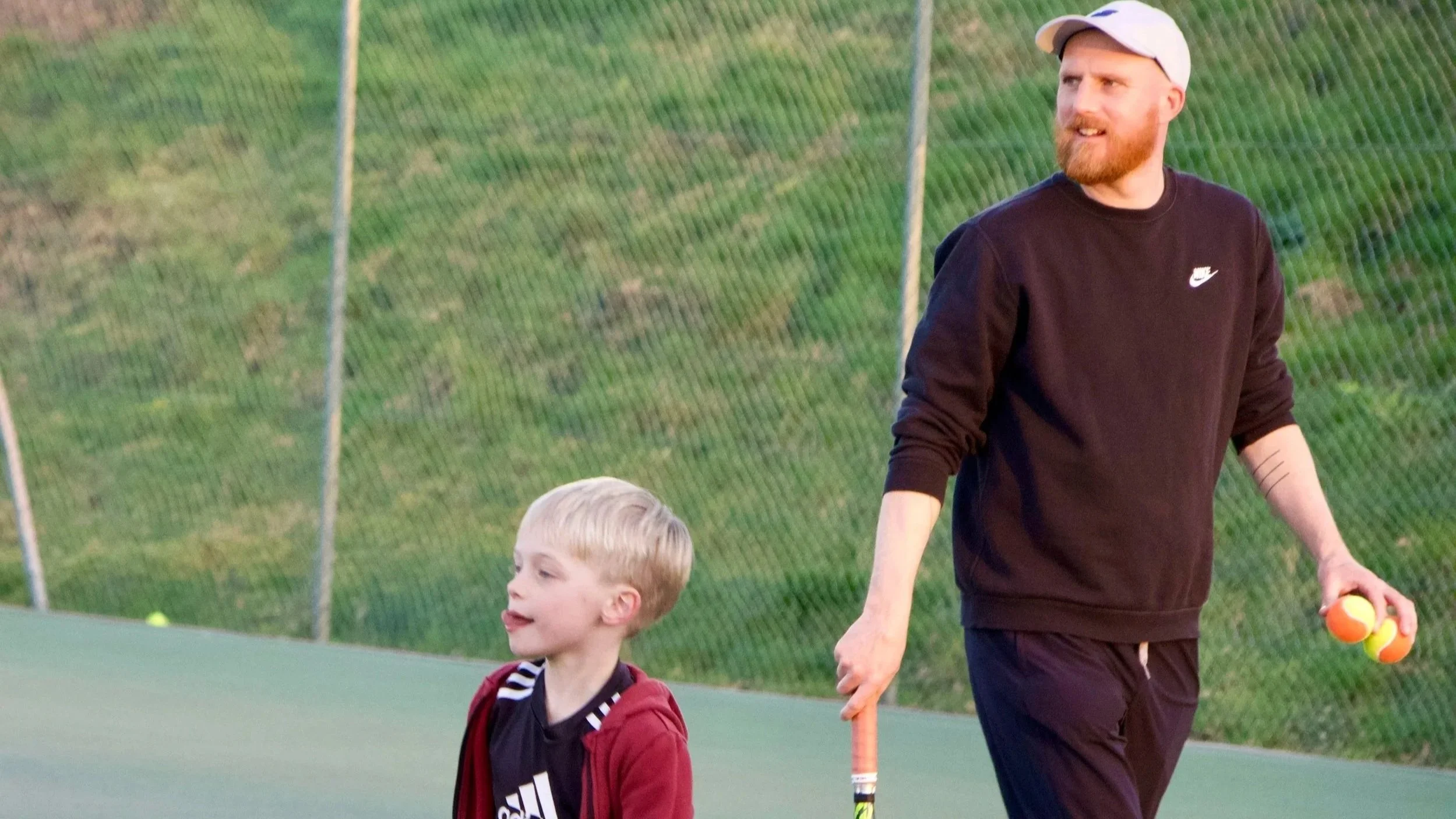 An adult man and a young boy walking on a tennis court. The man is holding several tennis balls in his left hand and a tennis racket in his right hand. The background shows a chain-link fence and grassy hill.