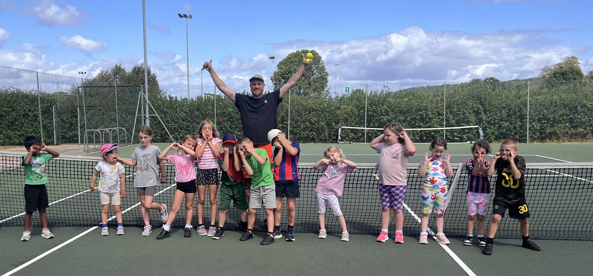 A group of children and an adult man standing on a tennis court behind the net. The man is holding two tennis balls and smiling, with his arms raised. The children are making playful faces and poses, dressed casually, with some also holding tennis balls.