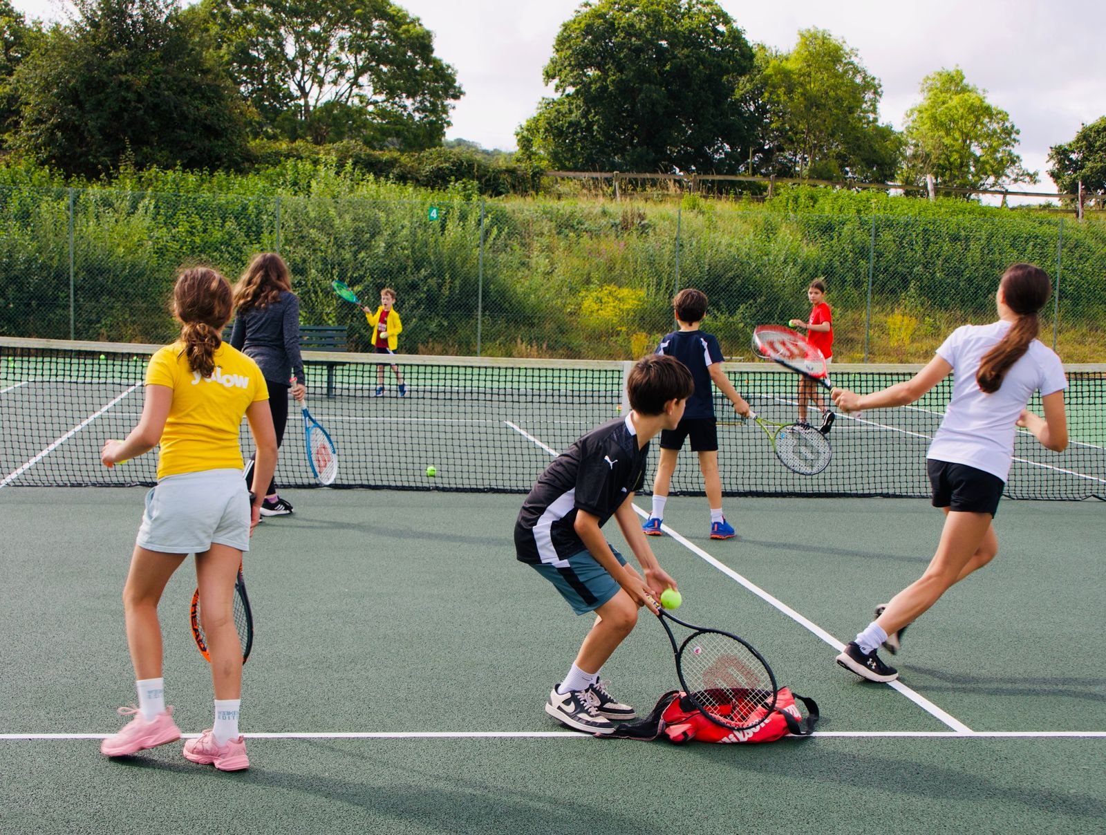 Children playing tennis on an outdoor court with a net, surrounded by green trees and a hillside.