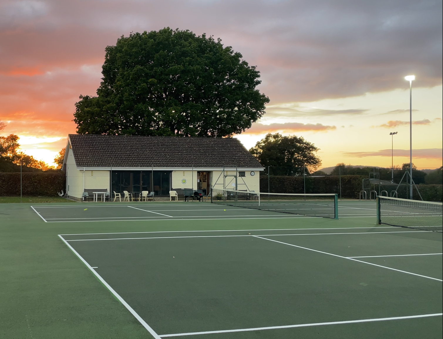 Empty tennis courts at sunset with a building and large trees in the background.