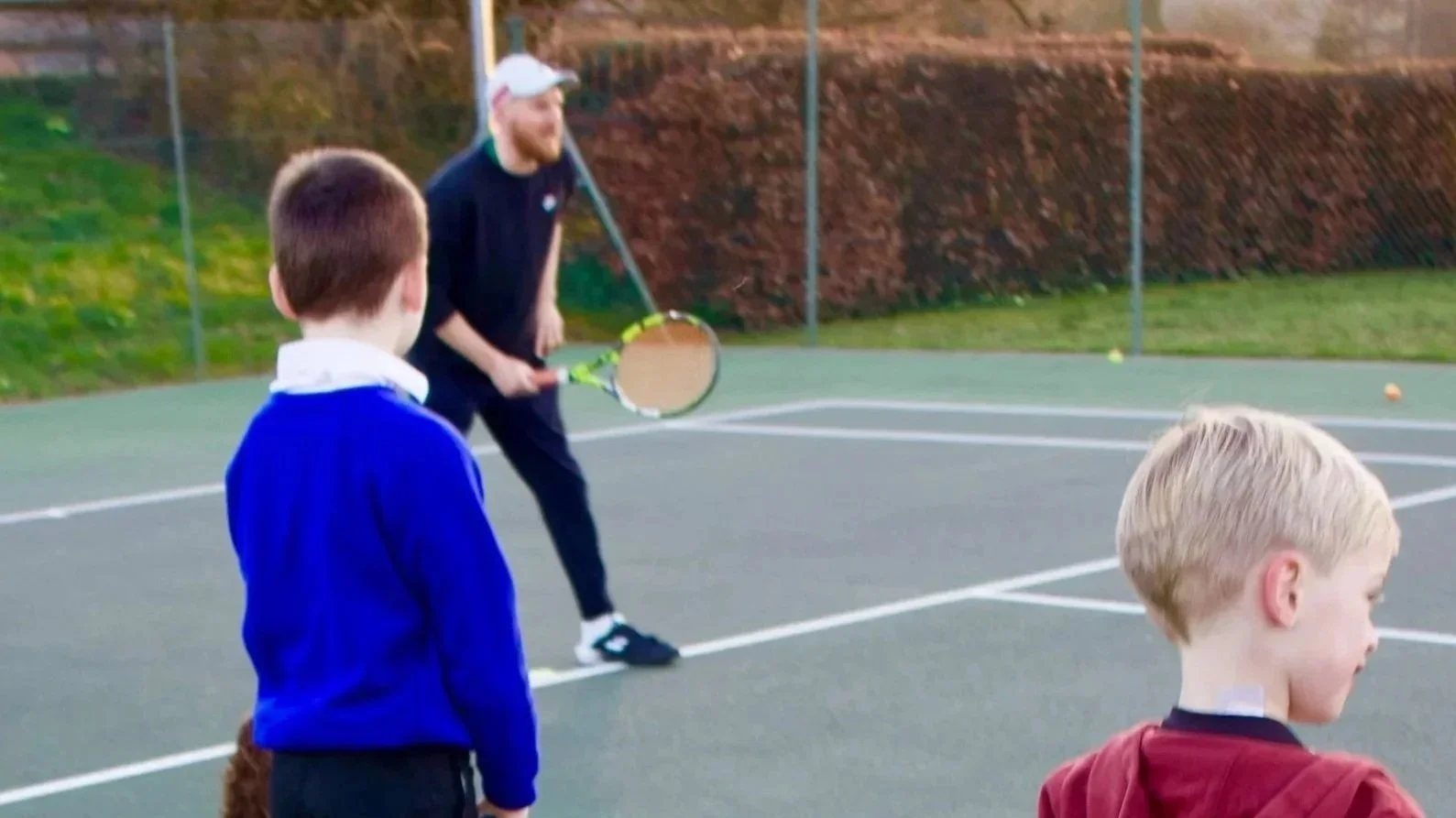 Two children and one adult playing tennis on an outdoor court during the daytime.