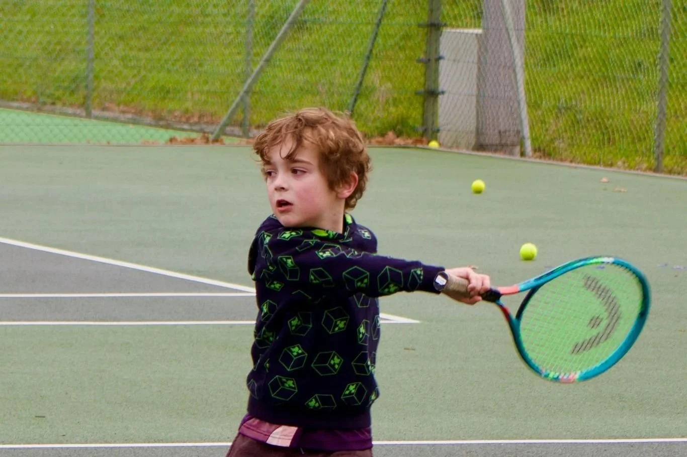A young boy with curly brown hair playing tennis on an outdoor court. He is holding a tennis racket and appears to be in motion, with tennis balls in the background. The court is surrounded by a chain-link fence and grassy area.