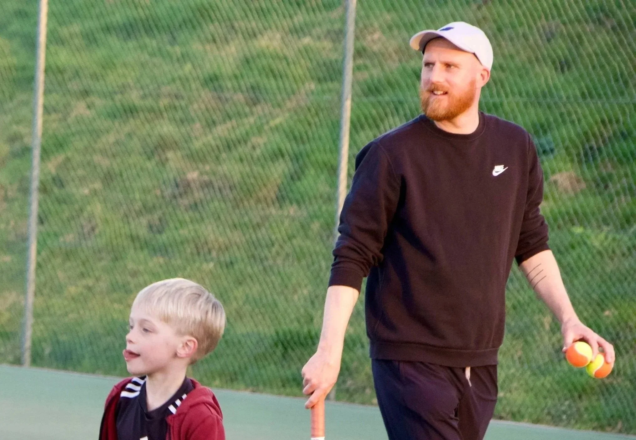 A man with a beard and a white cap holding two tennis balls and a tennis racket walking on a tennis court with a young boy in a red hoodie nearby.