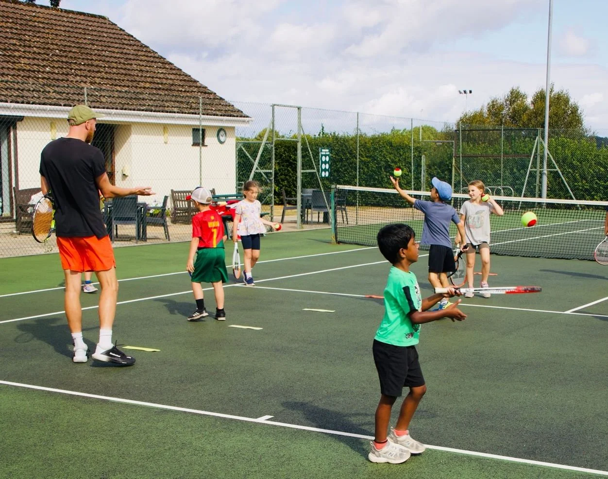 Children playing tennis on an outdoor court with a coach, some children holding tennis balls, and others preparing to serve or return shots. The court is surrounded by a fence and a building is visible in the background.