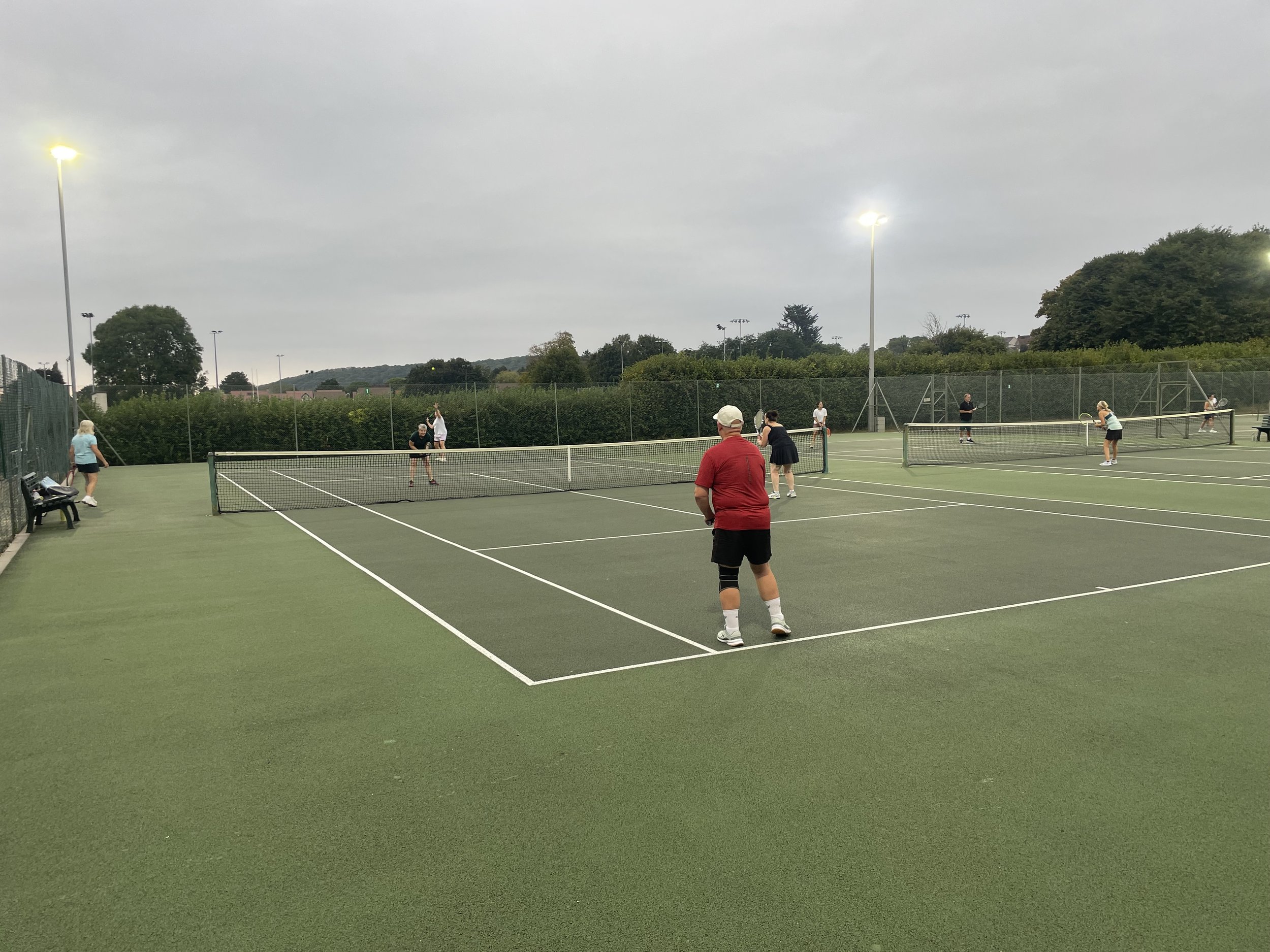 People playing doubles tennis on outdoor courts under cloudy sky during evening.