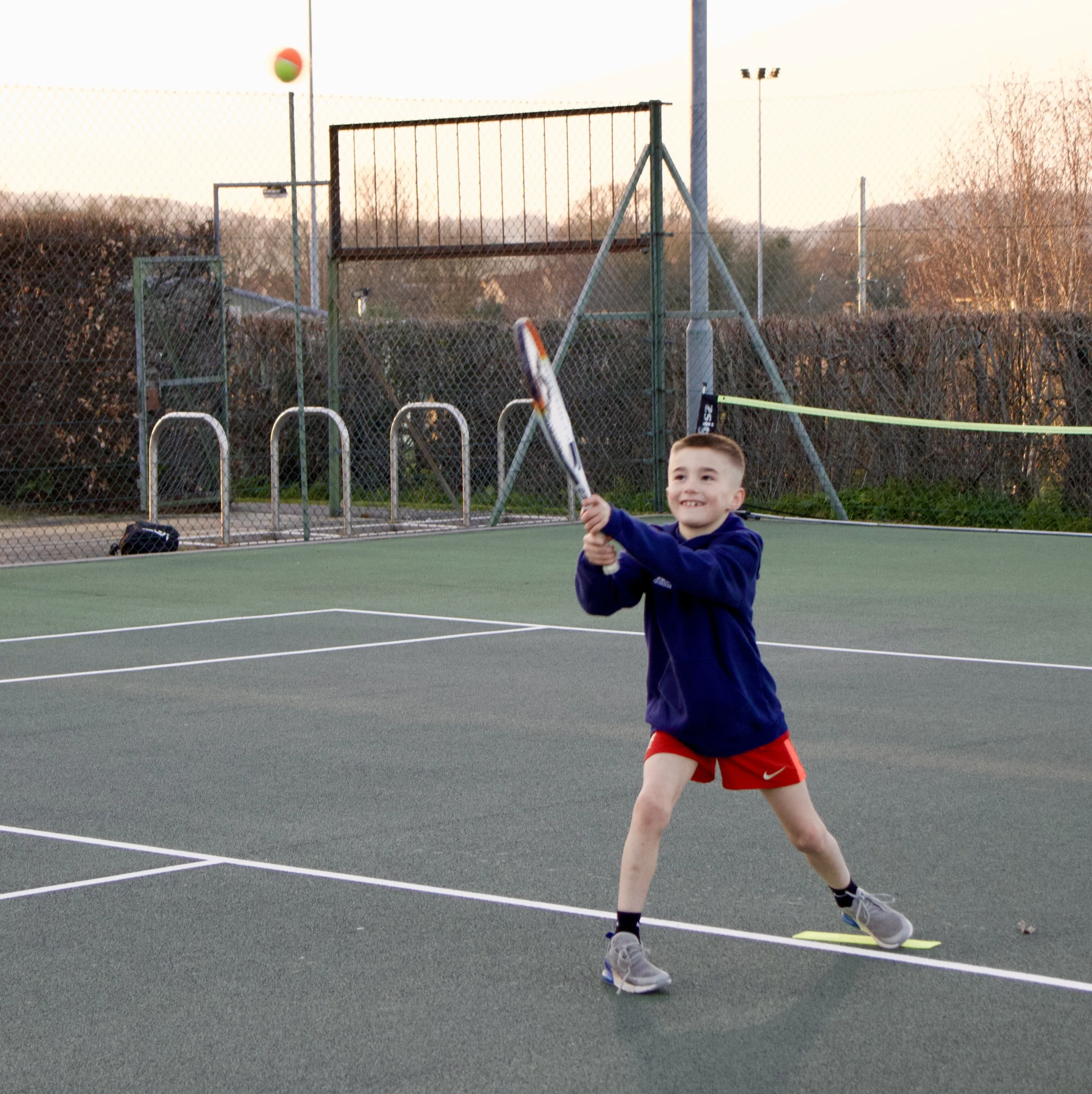 Young boy playing tennis on an outdoor court with a happy smile, wearing a navy hoodie, red shorts, and gray sneakers, holding a tennis racket, during evening with trees and fencing in background.