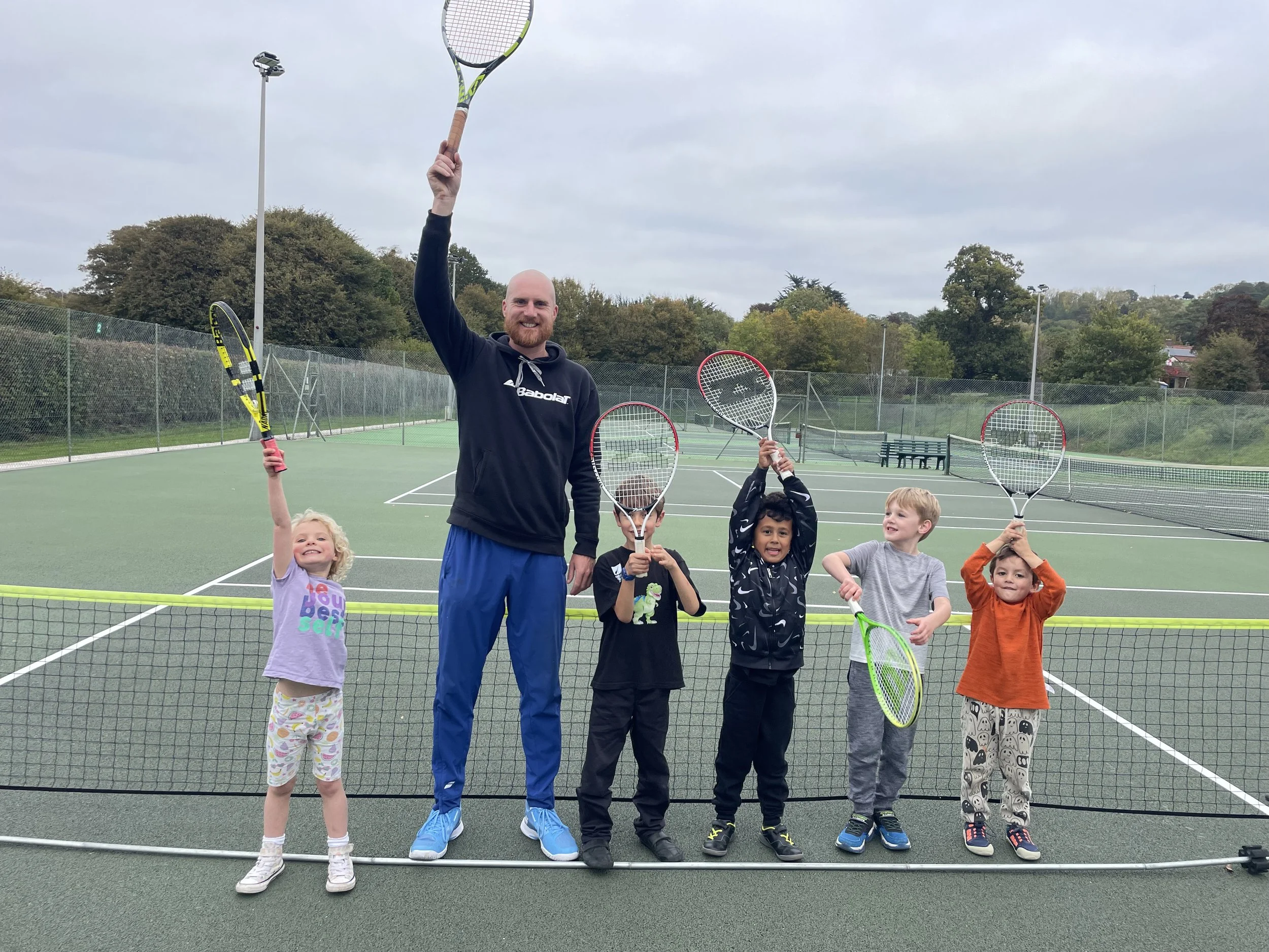 A man and five children, holding tennis rackets, stand on a tennis court. The man is smiling and holding a tennis racket high above his head, while the children pose with their rackets, some raised and some at their sides. The court is outdoors with trees and a cloudy sky in the background.