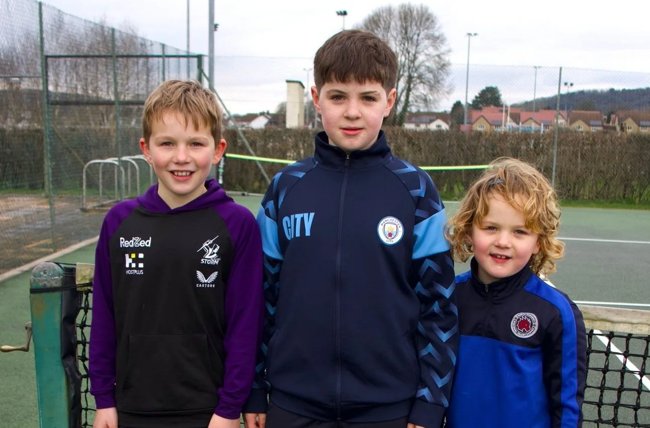 Three boys standing on a tennis court, wearing sports jackets and smiling at the camera.