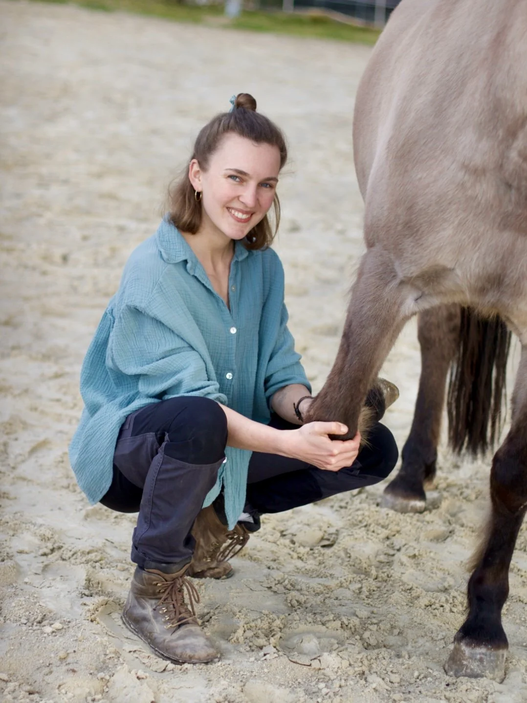 Eine lachende Frau kniet im Sand und hält das Vorderbein eines Pferdes.