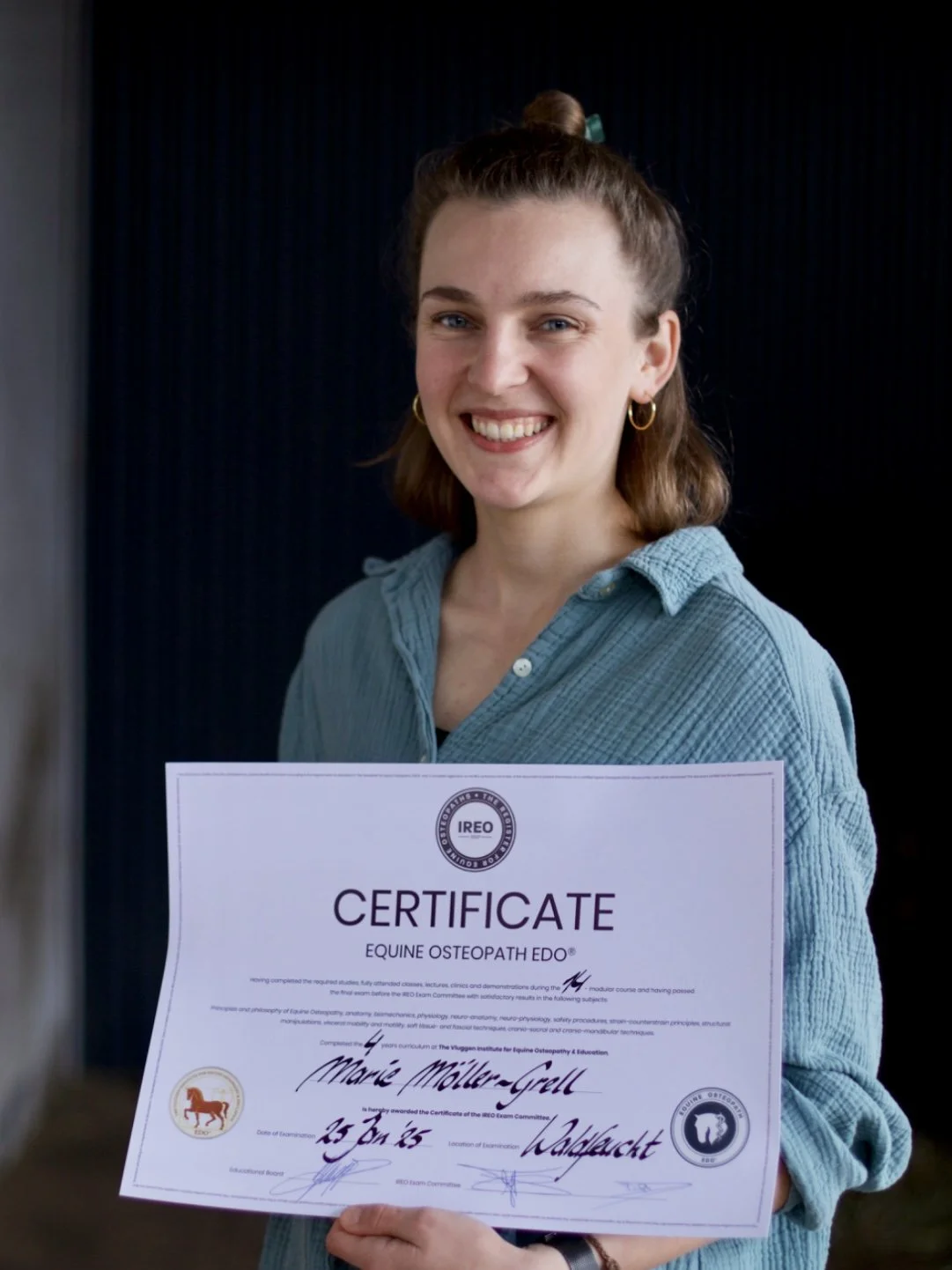 Eine lachende junge Frau hält ein Zertifikat für Equine Osteopathie. Sie trägt eine blaue Hemdbluse, hat braune Haare, Zöpfe im Haar und trägt goldene Ohrringe.