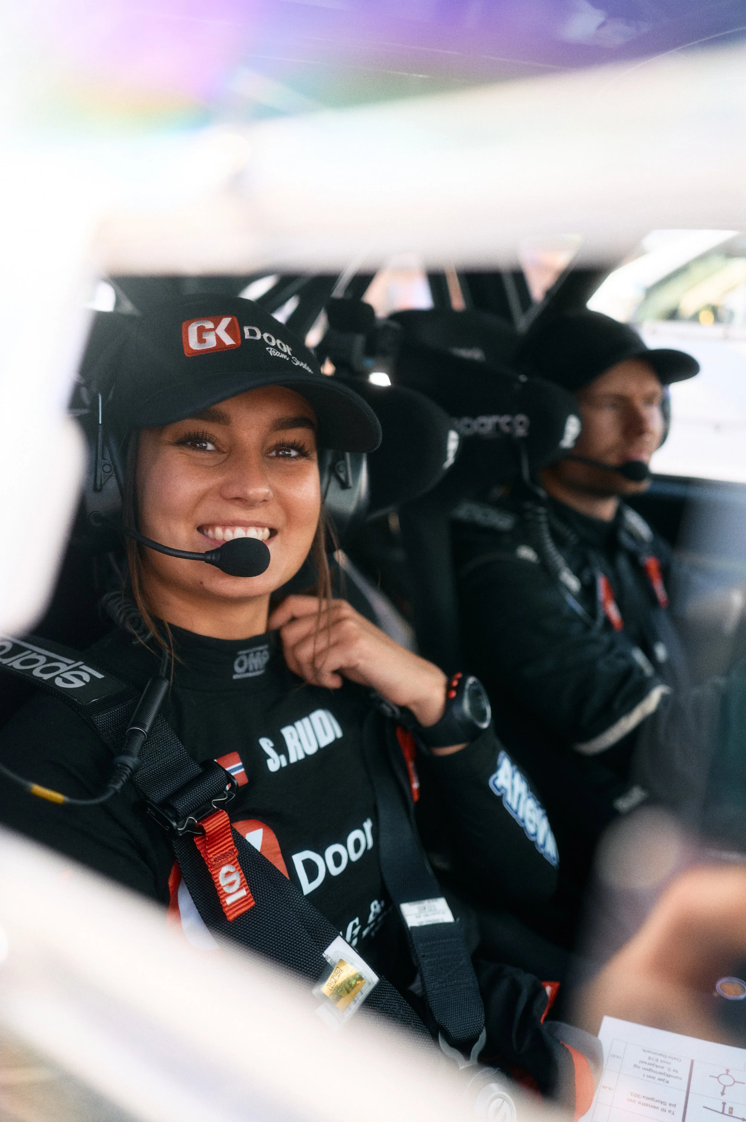 Two race car drivers in black racing suits and helmets with headsets, seated inside a race car, with a woman smiling at the camera.