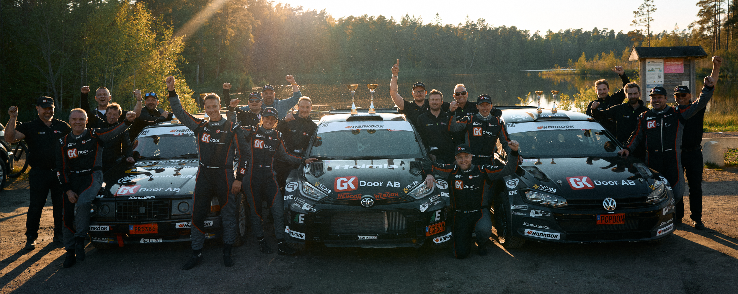 A racing team celebrating with three race cars, four trophies on the roof, and members cheering with clenched fists outdoors during sunset, near a lake and trees.