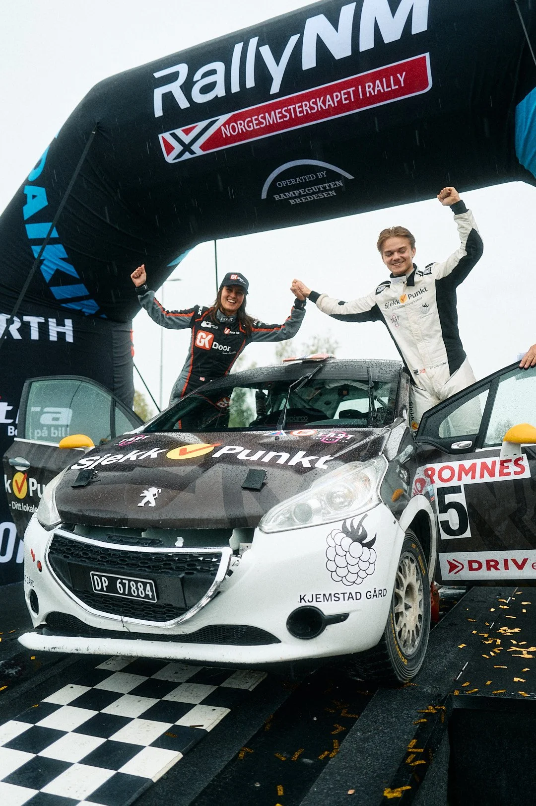 Two rally drivers celebrating on top of a race car under a banner that reads Rally NM, with rain falling and a checkered finish line beneath the car.