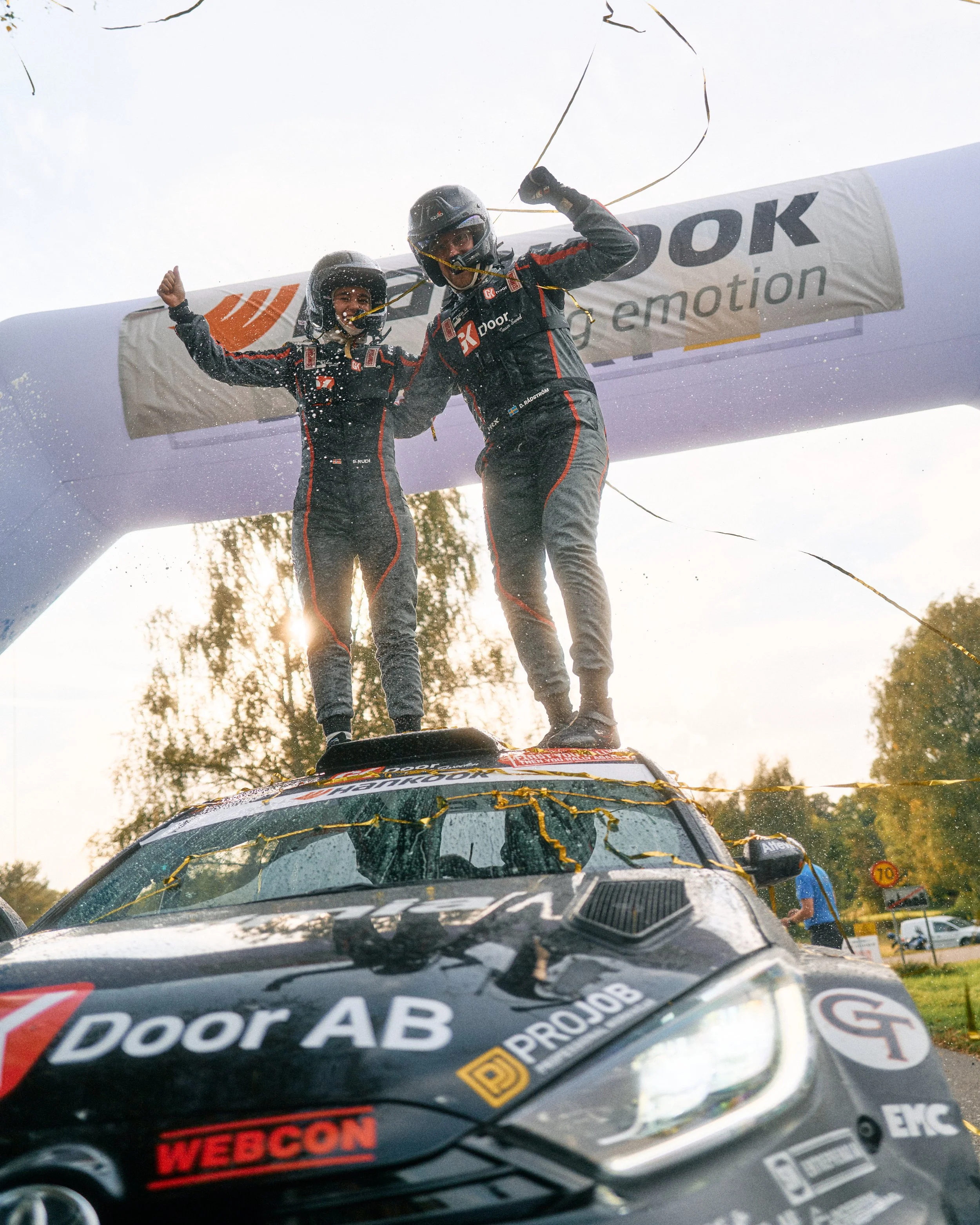 Two race car drivers in racing suits and helmets standing on top of a racing car celebrating victory beneath a finish line arch.