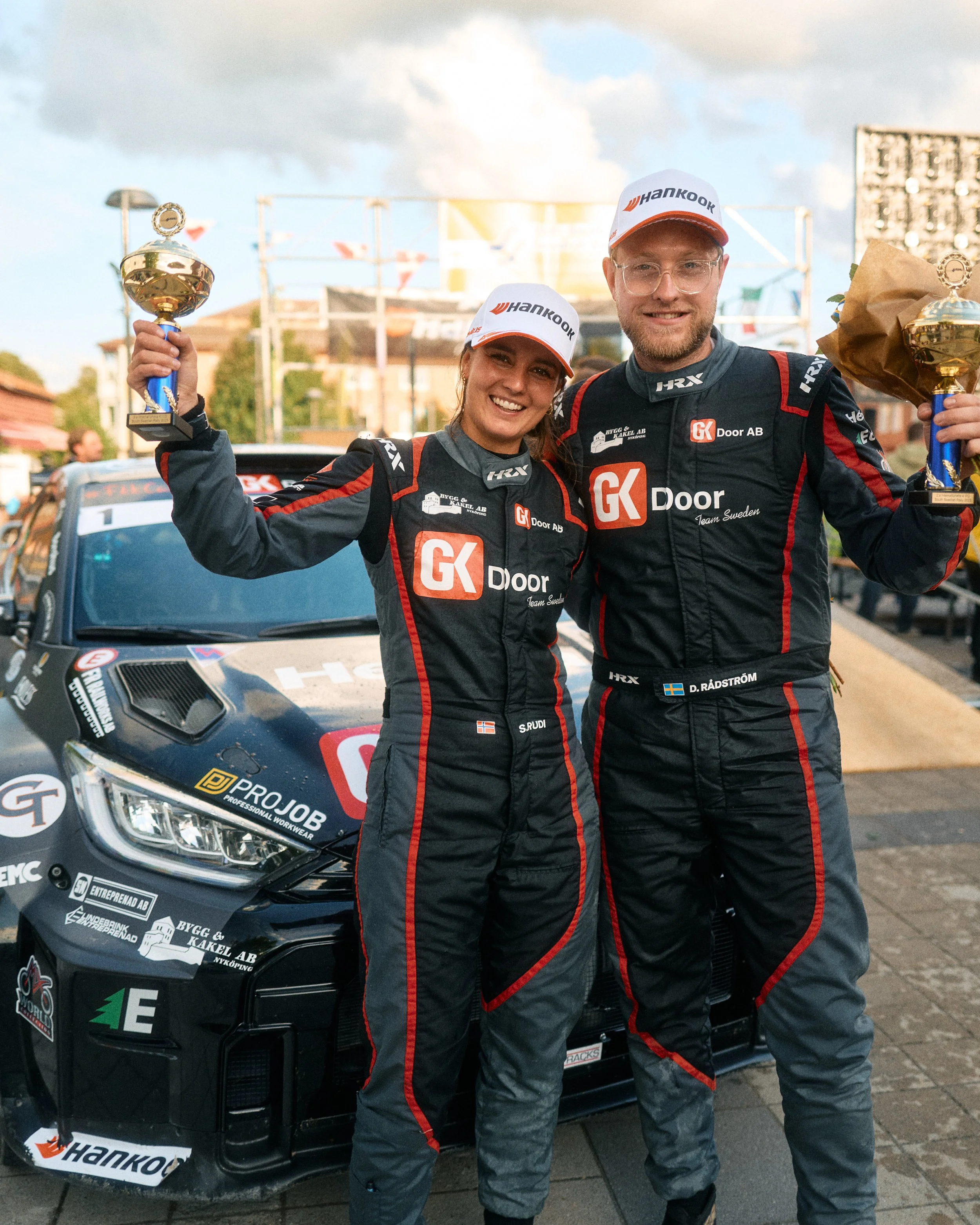 Two race car drivers celebrating with trophies and smiling in front of a race car, during a rally event.
