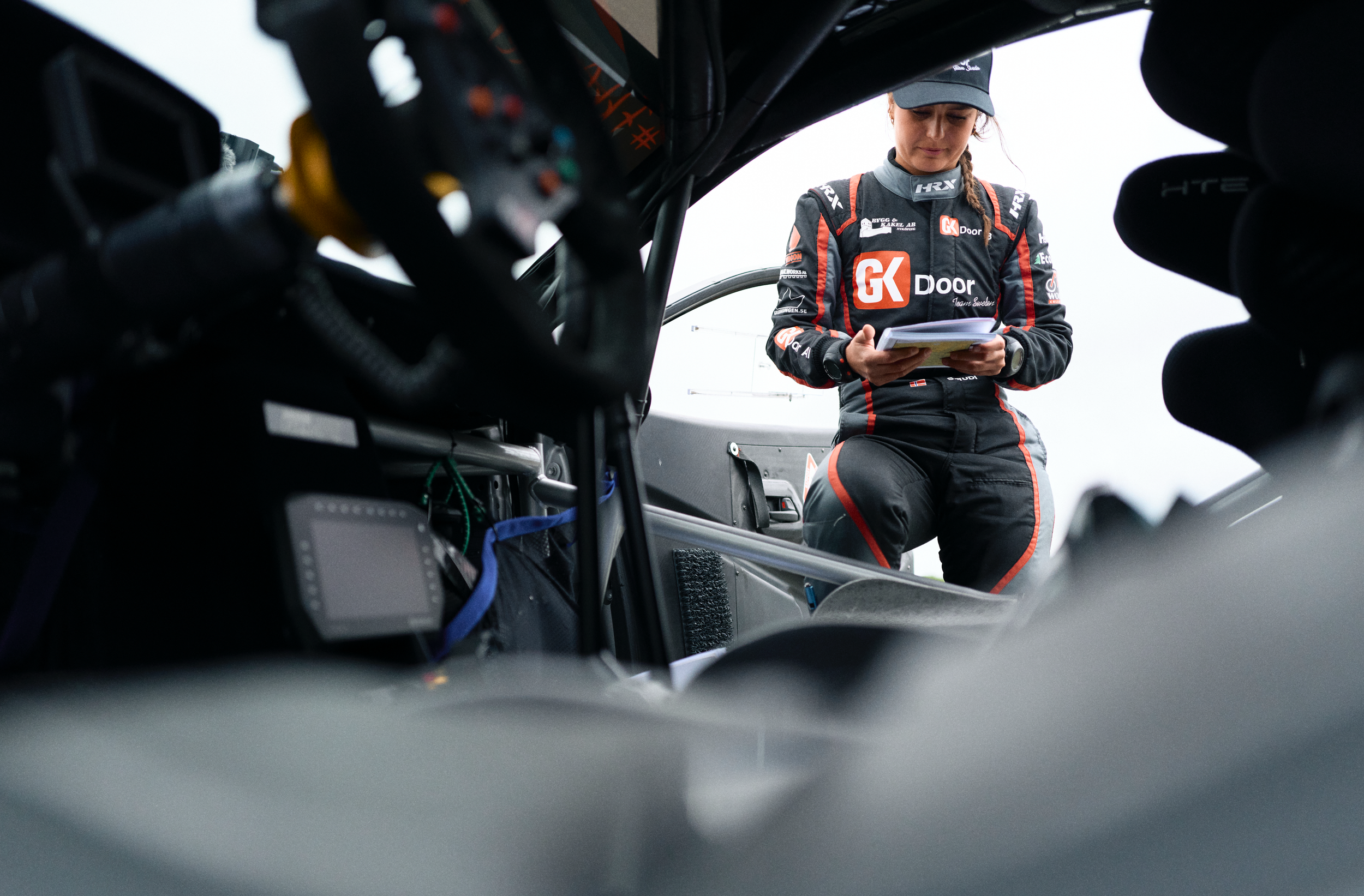 A female race car driver standing inside a race car, looking at a clipboard, wearing a black racing suit with red and white sponsorship logos, and a gray cap.