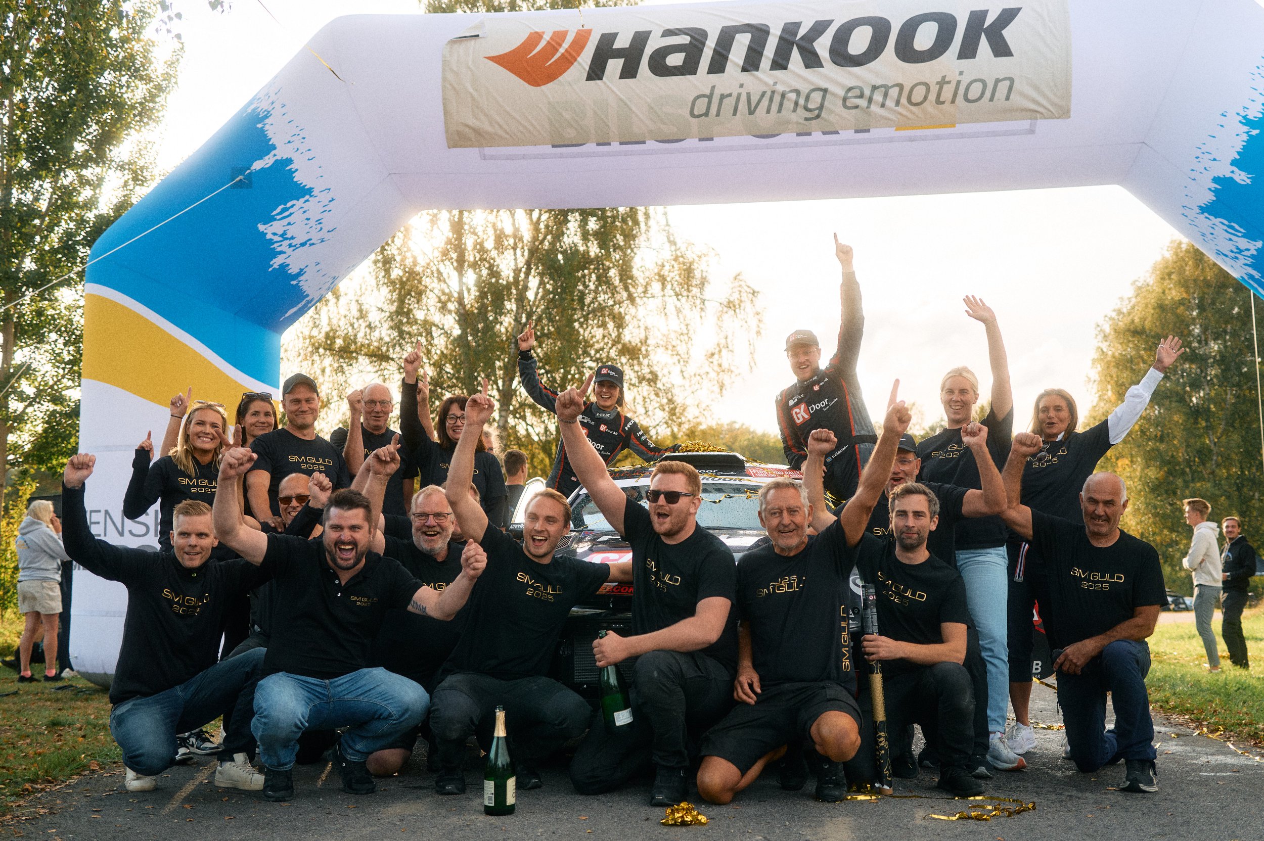 Celebrating a victory with a team of rally car drivers and crew at an outdoor event, standing and kneeling under a large inflatable arch with the Hankook logo. The team is in black shirts, some holding champagne bottles, with two drivers standing on the car celebrating. The background has trees and a few people observing.