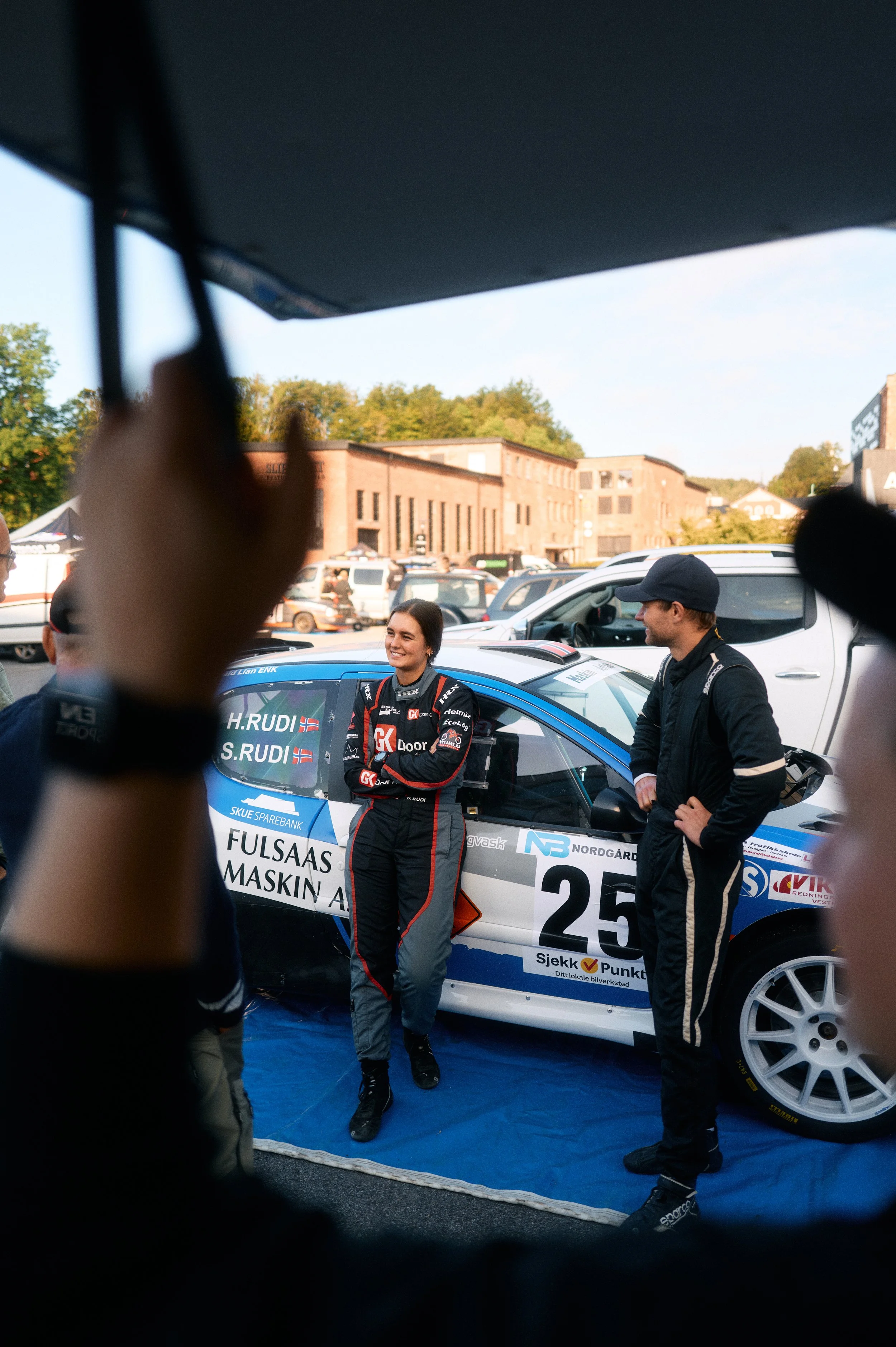 A female rally car driver standing beside her race car, smiling and talking to a man, at a racing event in a parking lot with vehicles and buildings in the background.